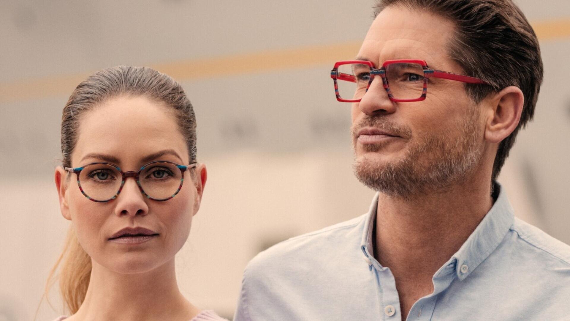 A woman and a man stand side by side, both wearing eyeglasses with bold, colorful frames. The woman has her hair pulled back and wears round glasses, while the man wears rectangular red glasses and a light blue shirt.
