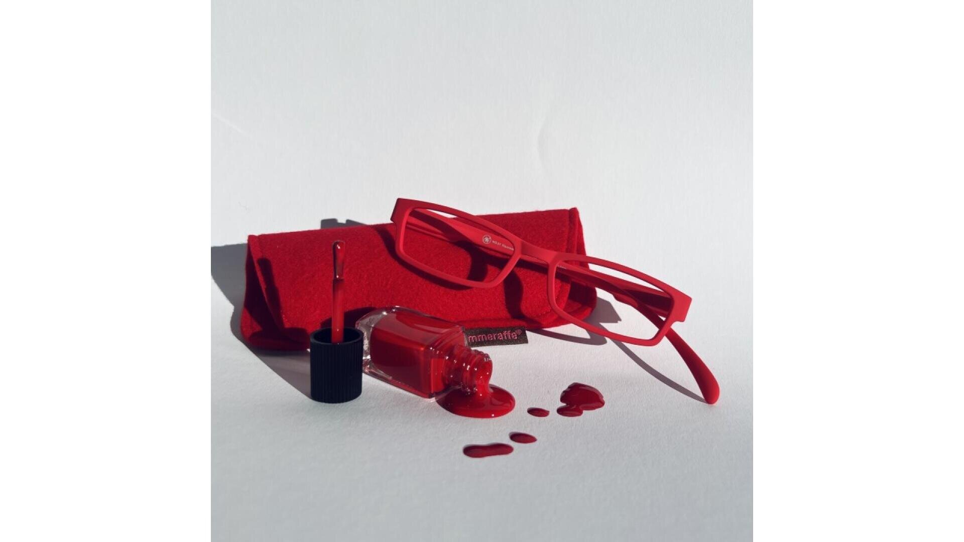Red eyeglasses and a matching red case are placed on a white surface. In front of them, a bottle of red nail polish lies open and spilled, with drops of polish and the applicator brush visible.