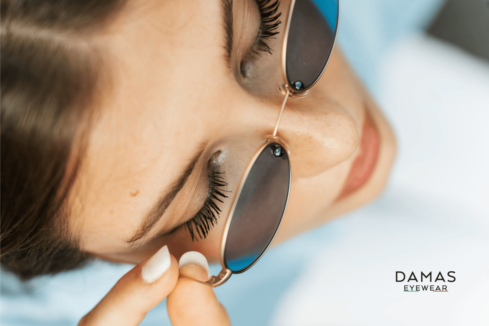 Close-up of a woman wearing sunglasses with golden rims and adjusting them with her hand. Her long eyelashes and fair skin can be seen. The DAMAS EYEWEAR logo appears at the bottom right.