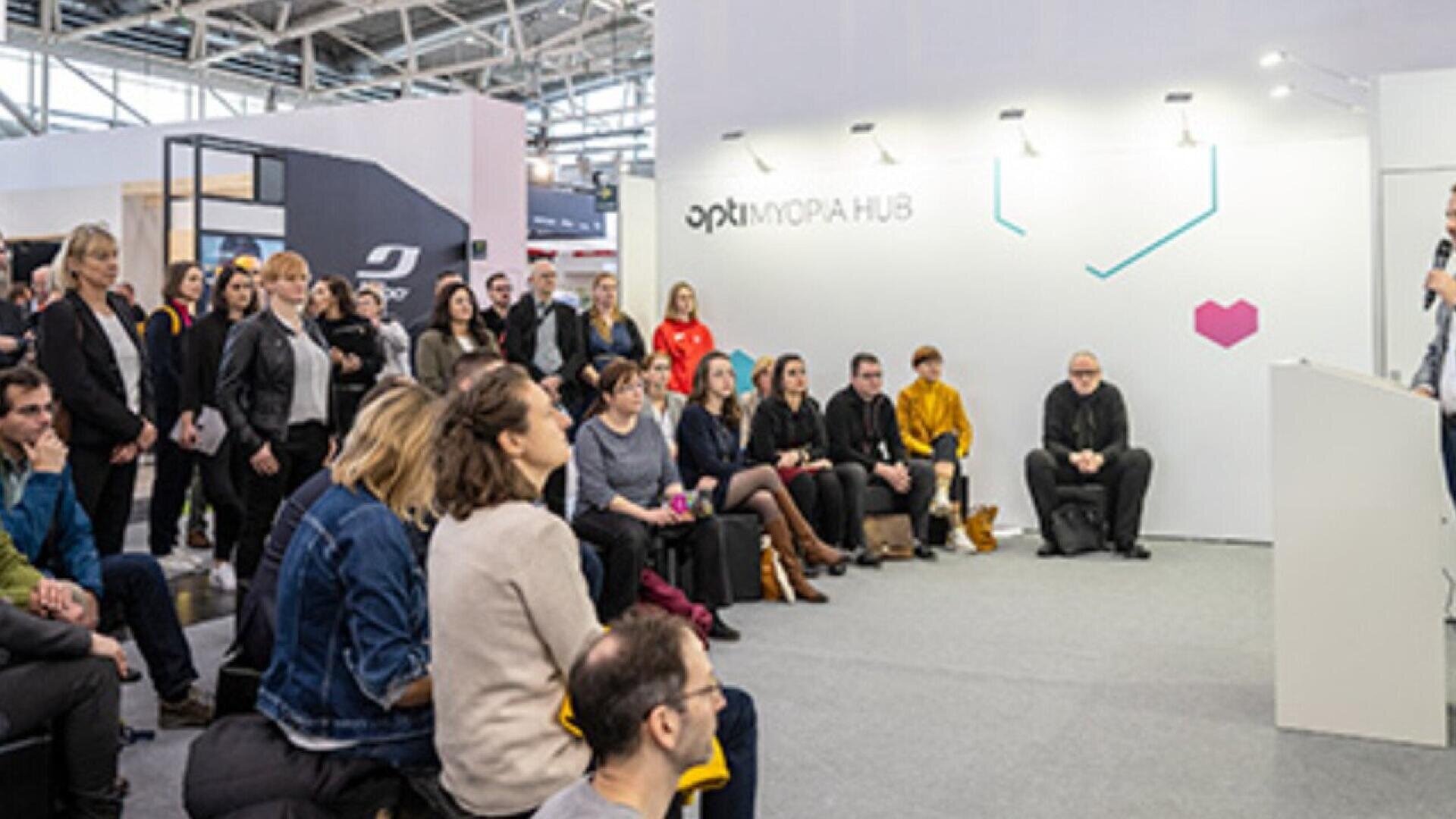 A group of people sits and listens attentively to a speaker at a modern indoor event or conference; the backdrop displays the text “OPTI MORPH HUB” and geometric decorations.