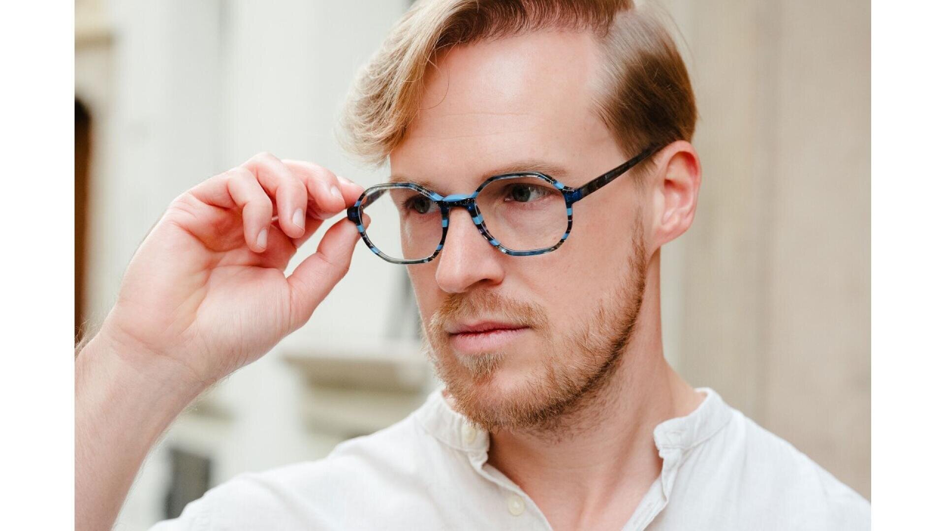 A man with light hair and a short beard wears a cream-colored shirt and blue-patterned eyeglasses, looking thoughtfully to the side while adjusting his glasses with his right hand.