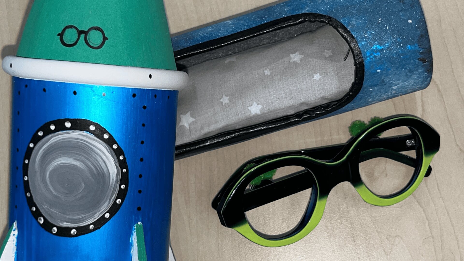 Eine blau-grüne Spielzeugrakete mit aufgemalter Brille steht neben einem passenden Brillenetui in Raketenform und einer schwarz-grünen Plastikbrille auf einer hellen Holzoberfläche.