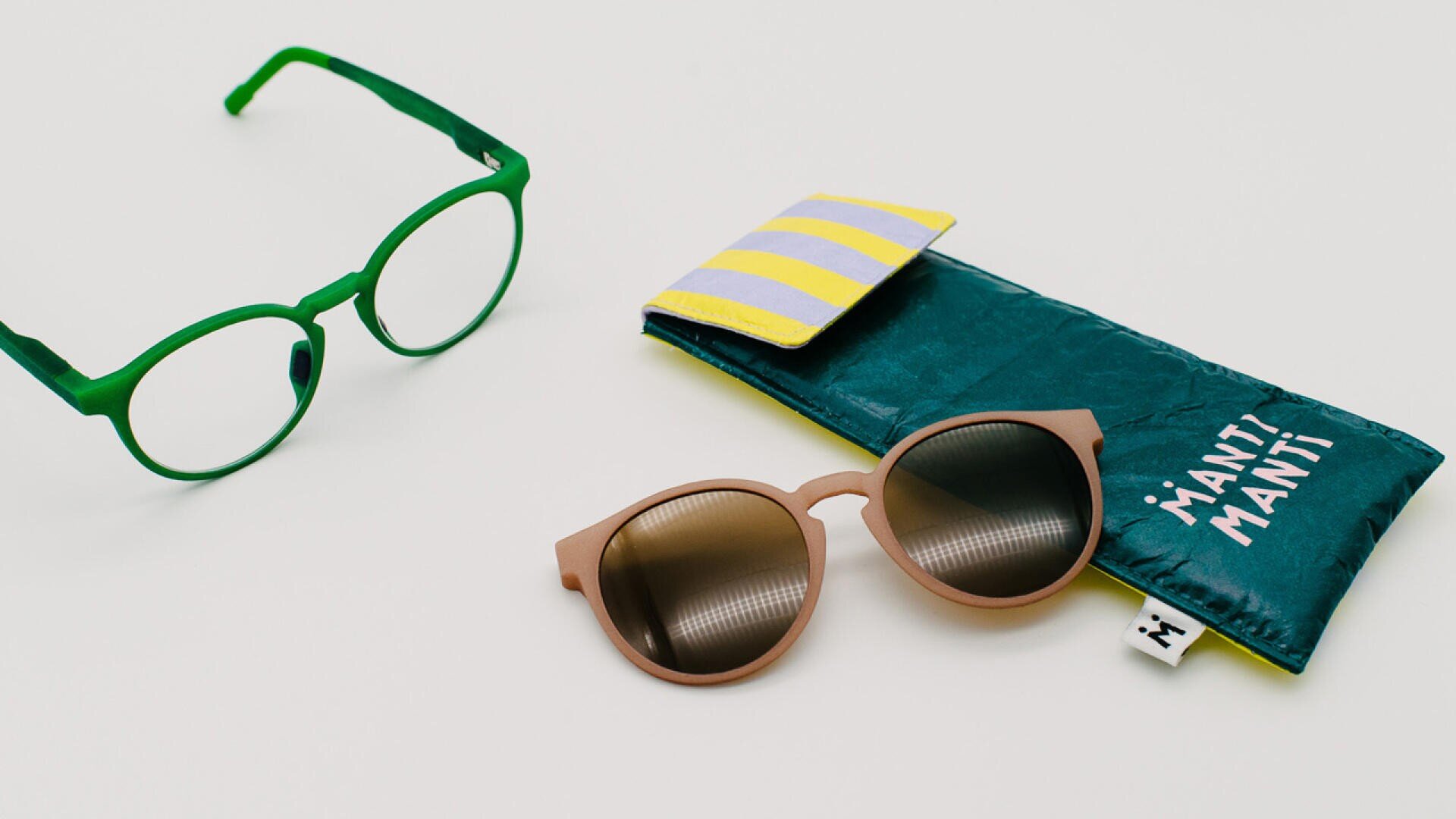 Eine grüne Brille, eine braune Sonnenbrille und ein grünes Brillenetui mit gelben Streifen und weißem Text, alles auf einer weißen Oberfläche angeordnet.
