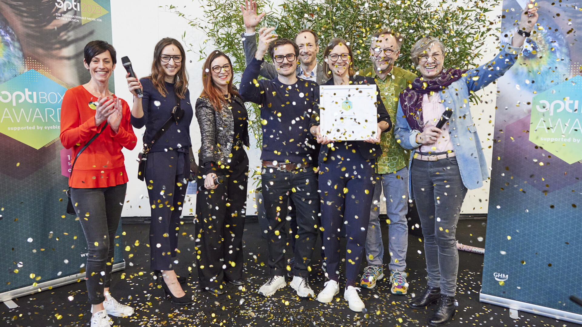 Eine Gruppe von Menschen feiert lächelnd auf der Bühne und hält eine Auszeichnung in den Händen, während goldenes Konfetti um sie herum herabregnet. Sie stehen zwischen zwei Bannern mit der Aufschrift „AWARD“.