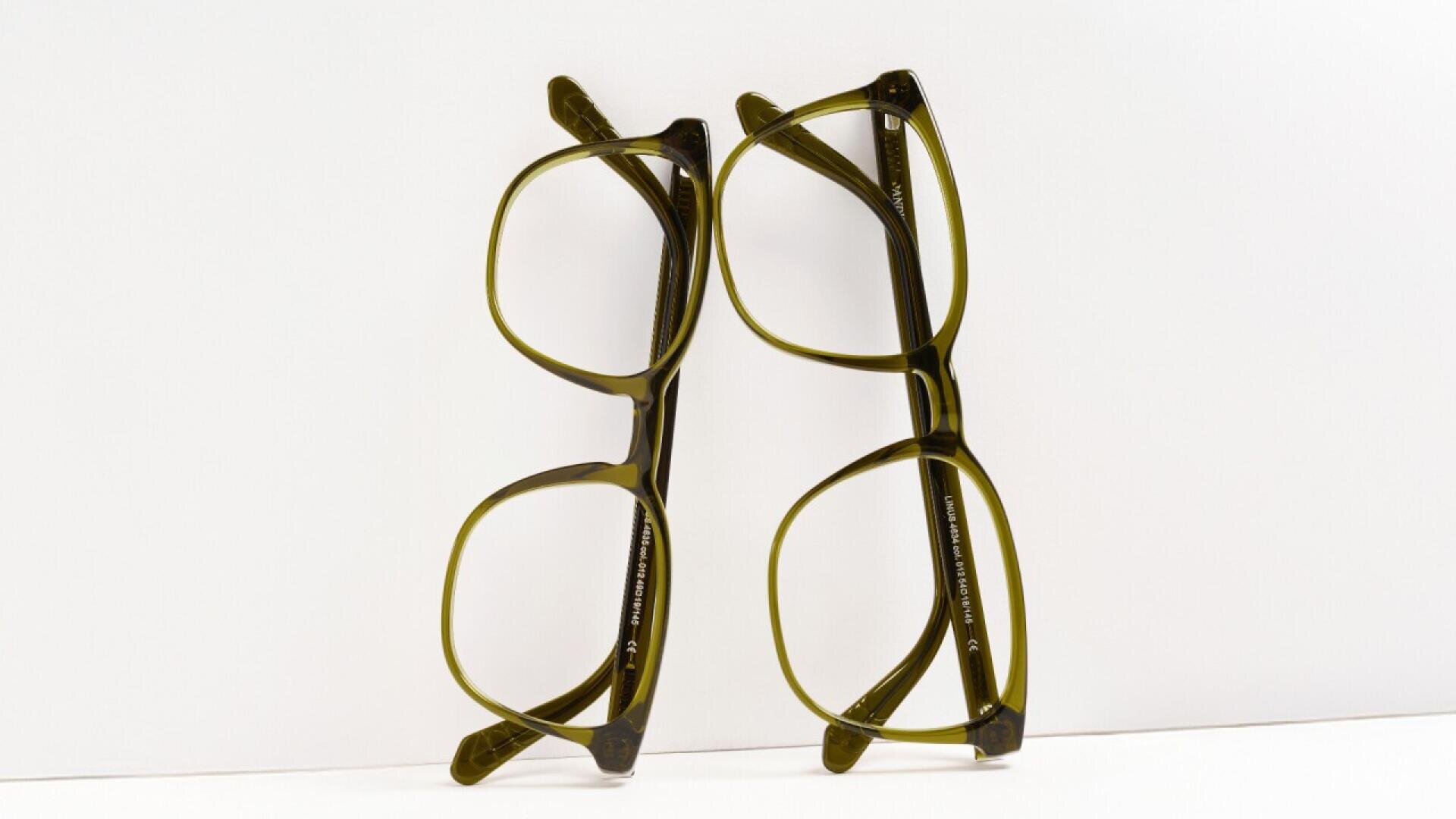 Two pairs of eyeglasses with greenish frames are standing upright side by side against a white background, leaning slightly towards each other.