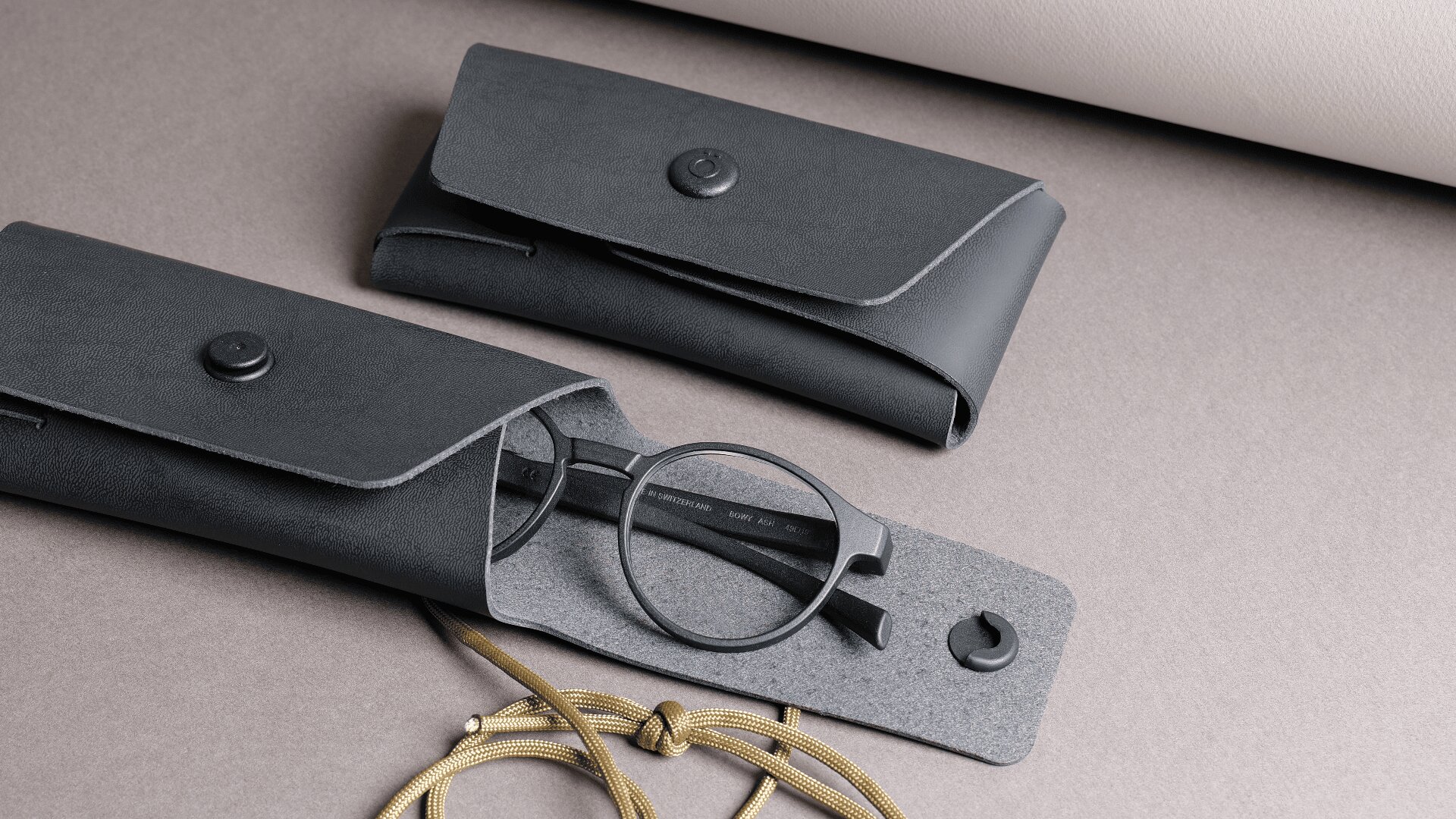 Eine schwarze Brille, teilweise in einem schwarzen Brillenetui, daneben ein weiteres geschlossenes Etui, alles auf einer grauen Fläche mit einer beigen Kordel und einer hellen Rolle im Hintergrund.