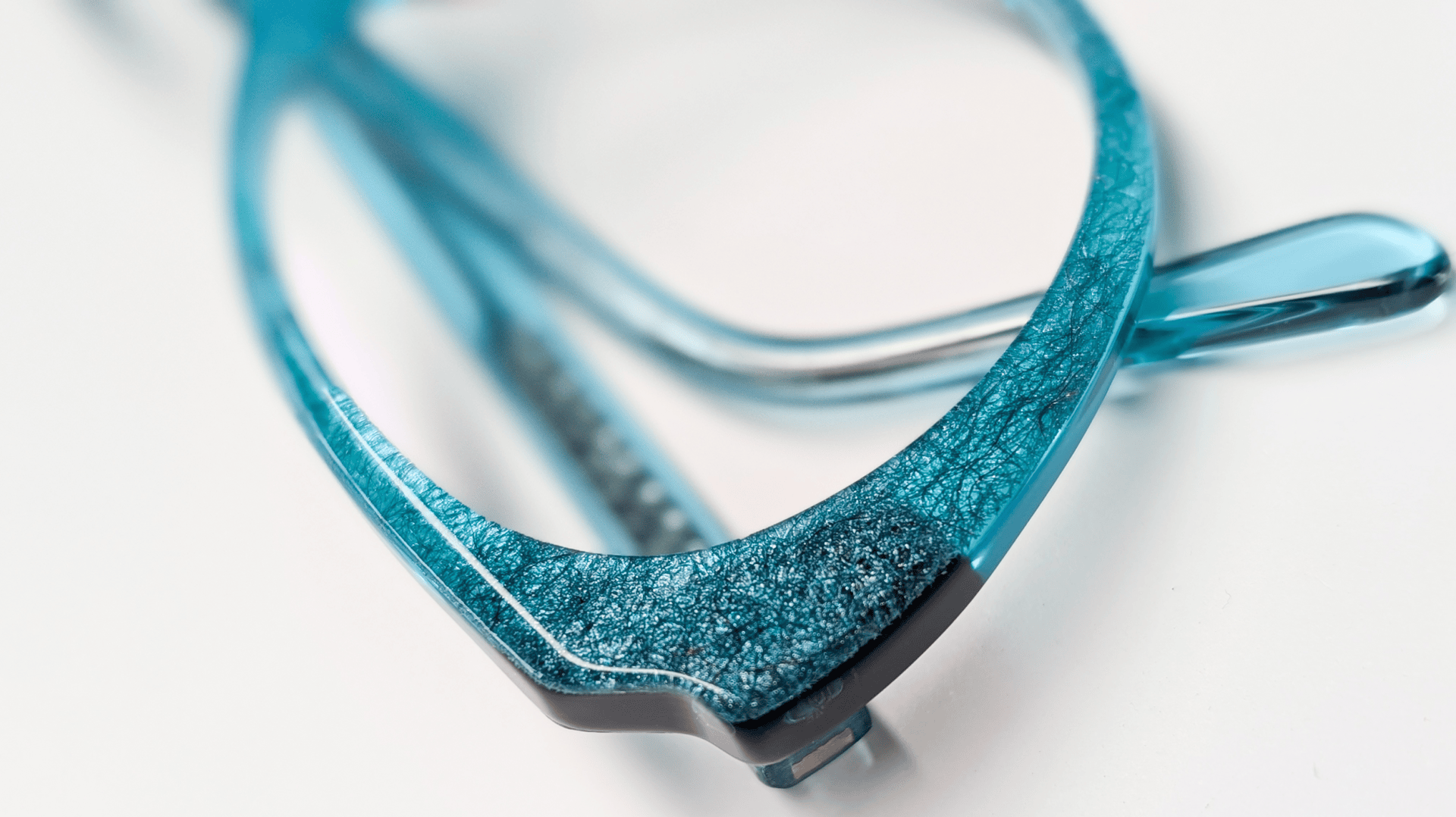 Nahaufnahme einer blauen Brille mit strukturierten, gekräuselten Details am Rahmen, die auf einer weißen Fläche liegt. Der Fokus liegt auf der vorderen Ecke des Brillengestells und hebt dessen Farbe und Design hervor.