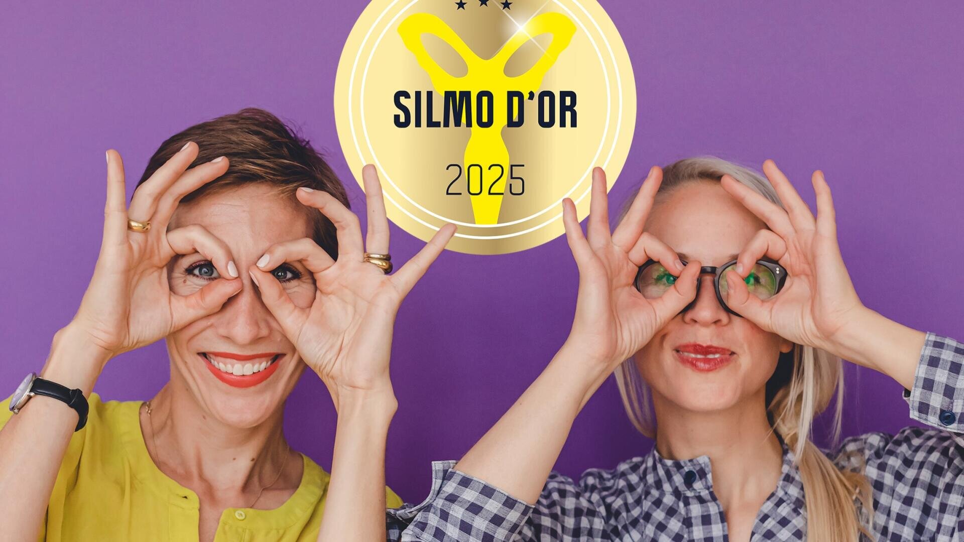 Two women stand in front of a purple background and form a pair of glasses with their hands over their eyes. A yellow SILMO DOR 2025 logo can be seen above them. Both are smiling and appear cheerful.