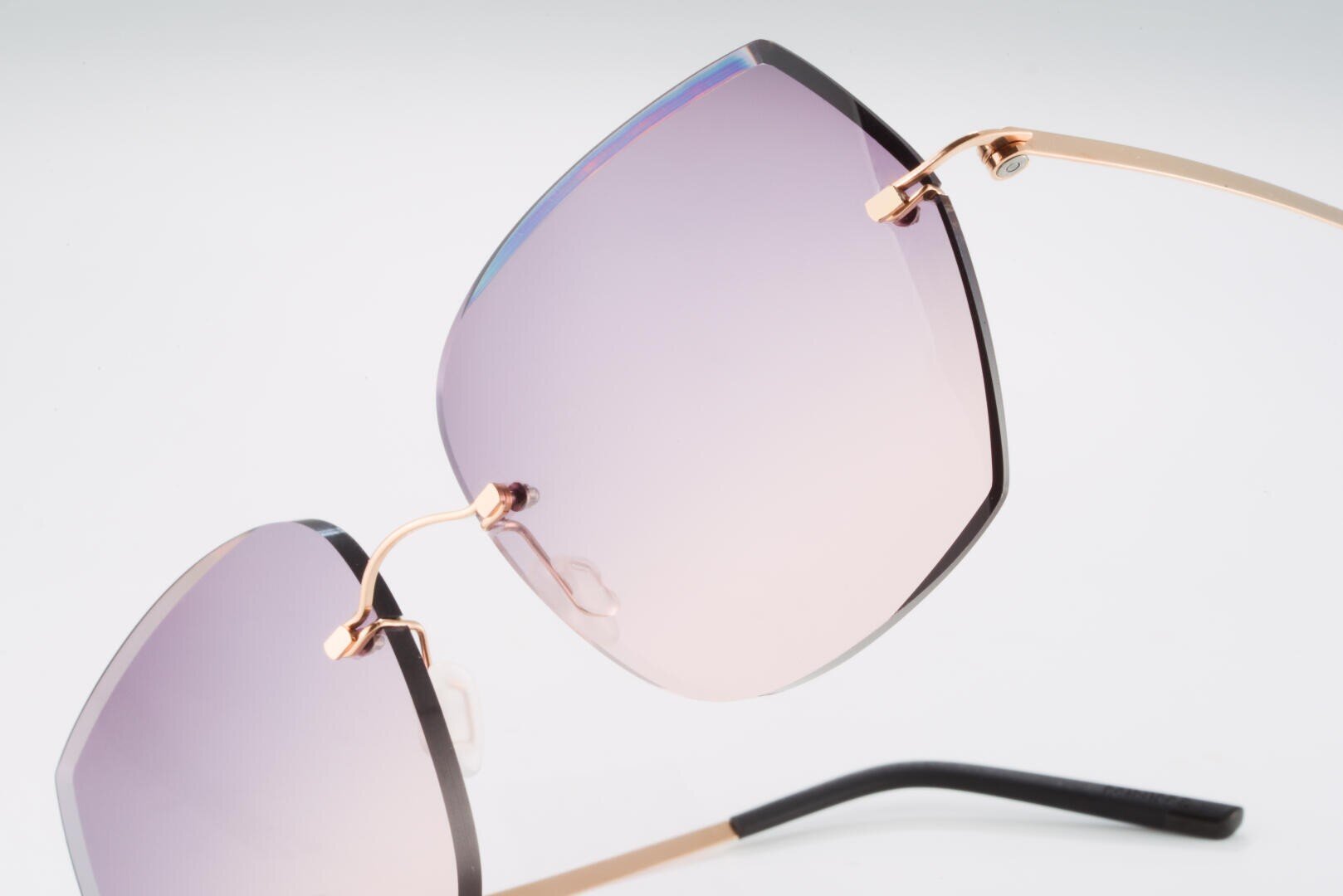 Nahaufnahme einer stilvollen Sonnenbrille mit großen, quadratischen, lilafarbenen Gläsern mit Farbverlauf und dünnen goldenen Metallrahmen vor einem schlichten weißen Hintergrund.