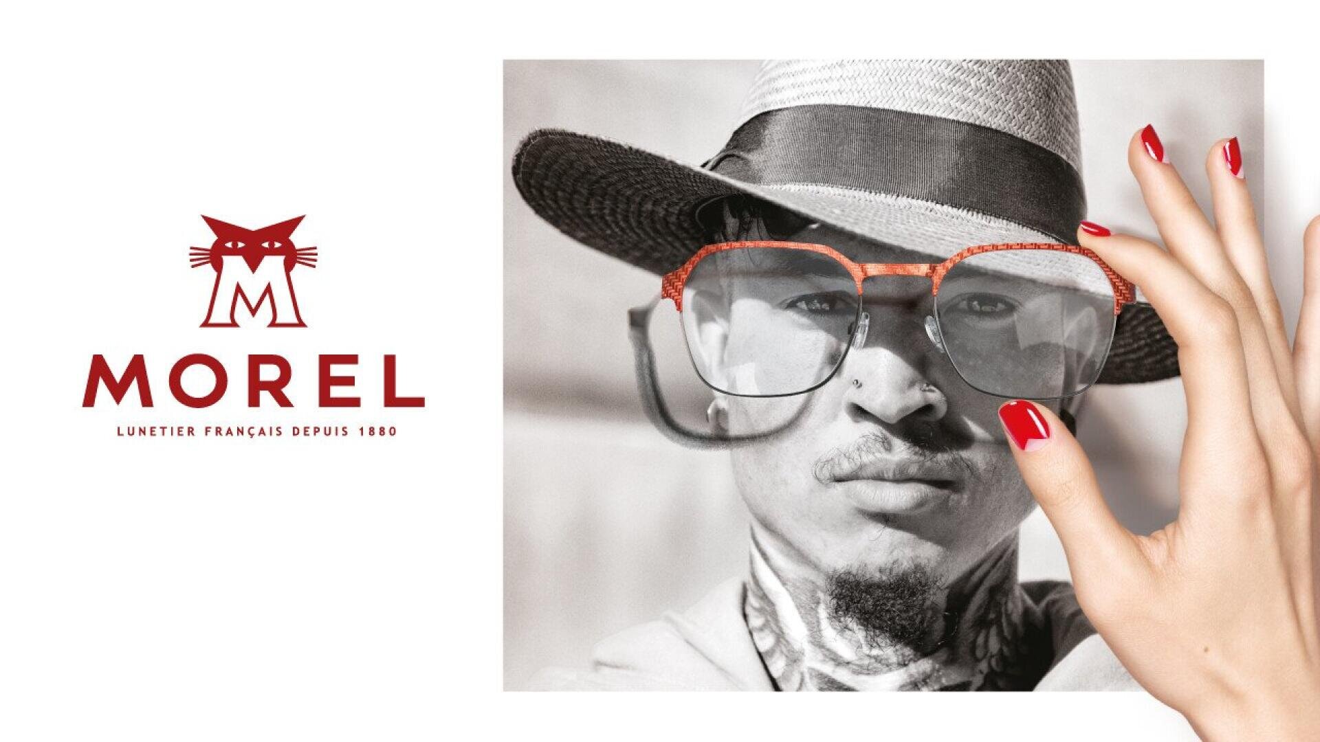 A person wearing a hat and oversized glasses is shown next to a red cat logo and the text MOREL Lunetier Français Depuis 1880. A hand with red nails holds the glasses in front of their face.