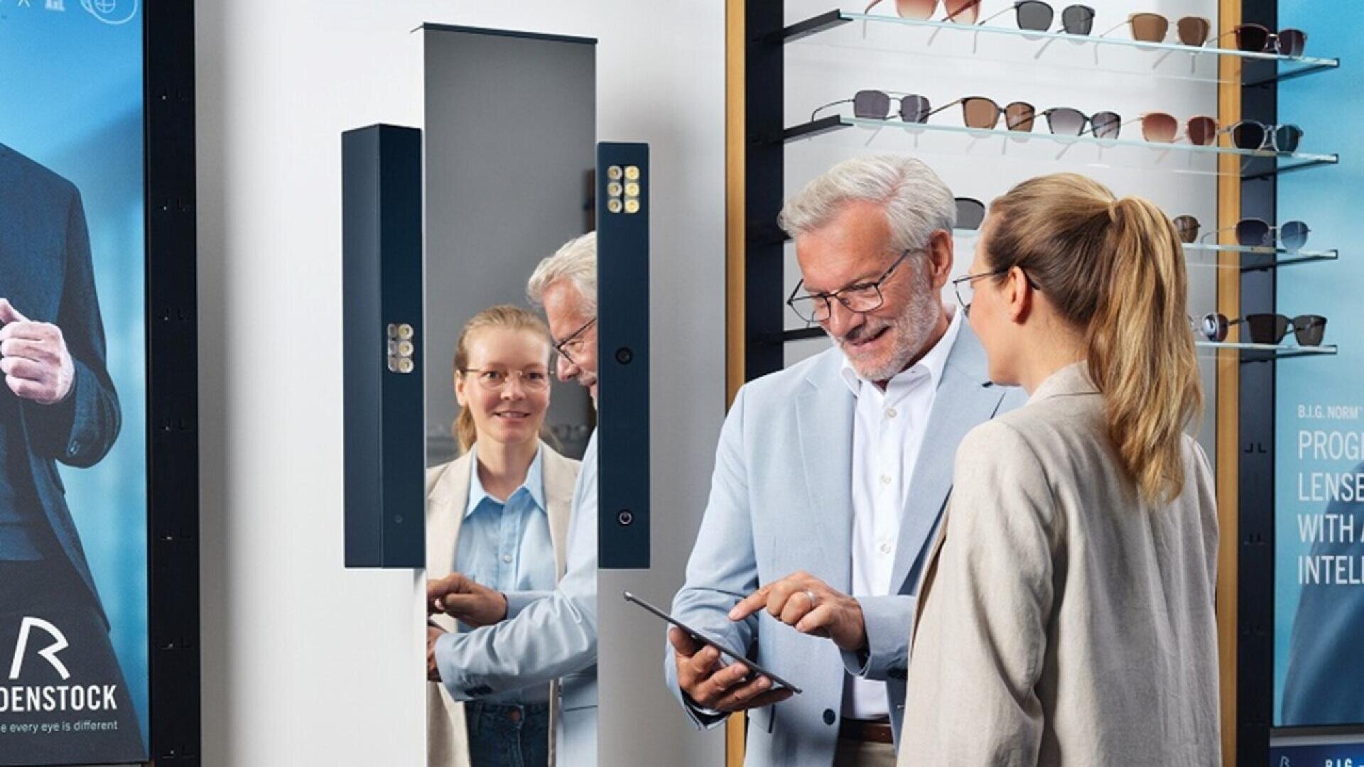 A smiling man in a light blue suit jacket holds a tablet and talks to a woman in a ponytail at an eyewear store, while the woman’s reflection appears in a nearby mirror. Glasses are displayed on shelves in the background.