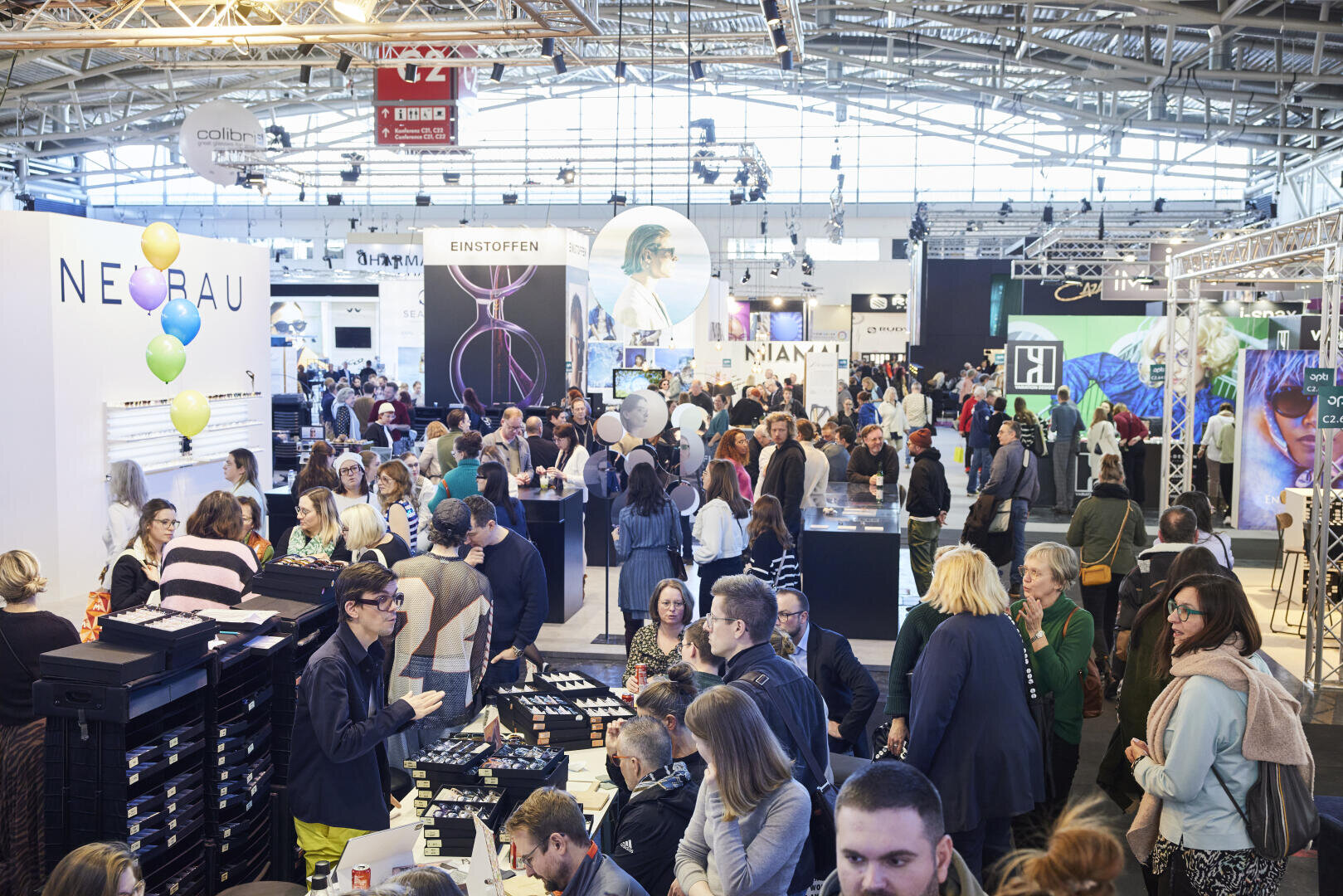 Eine geschäftige Messe- oder Ausstellungshalle ist voller Menschen, die unter hellem Licht miteinander interagieren, sich die Ausstellungsstücke ansehen und Produkte begutachten. Im Hintergrund sind verschiedene Firmenstände und Banner zu sehen.