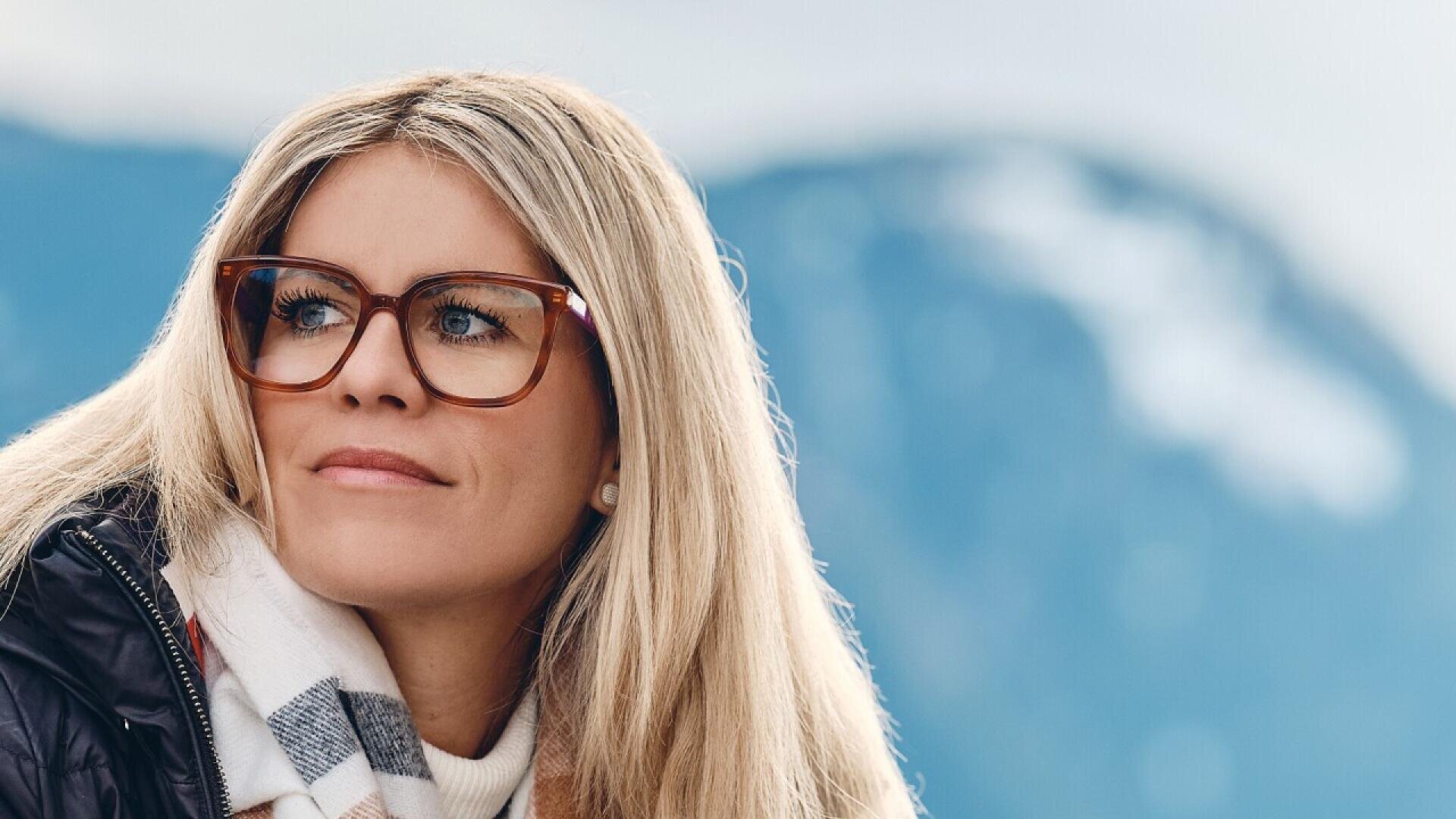 A woman with long blonde hair and brown glasses looks to the side, wearing a scarf and a jacket. Snowy mountains and a blurred sky are in the background.