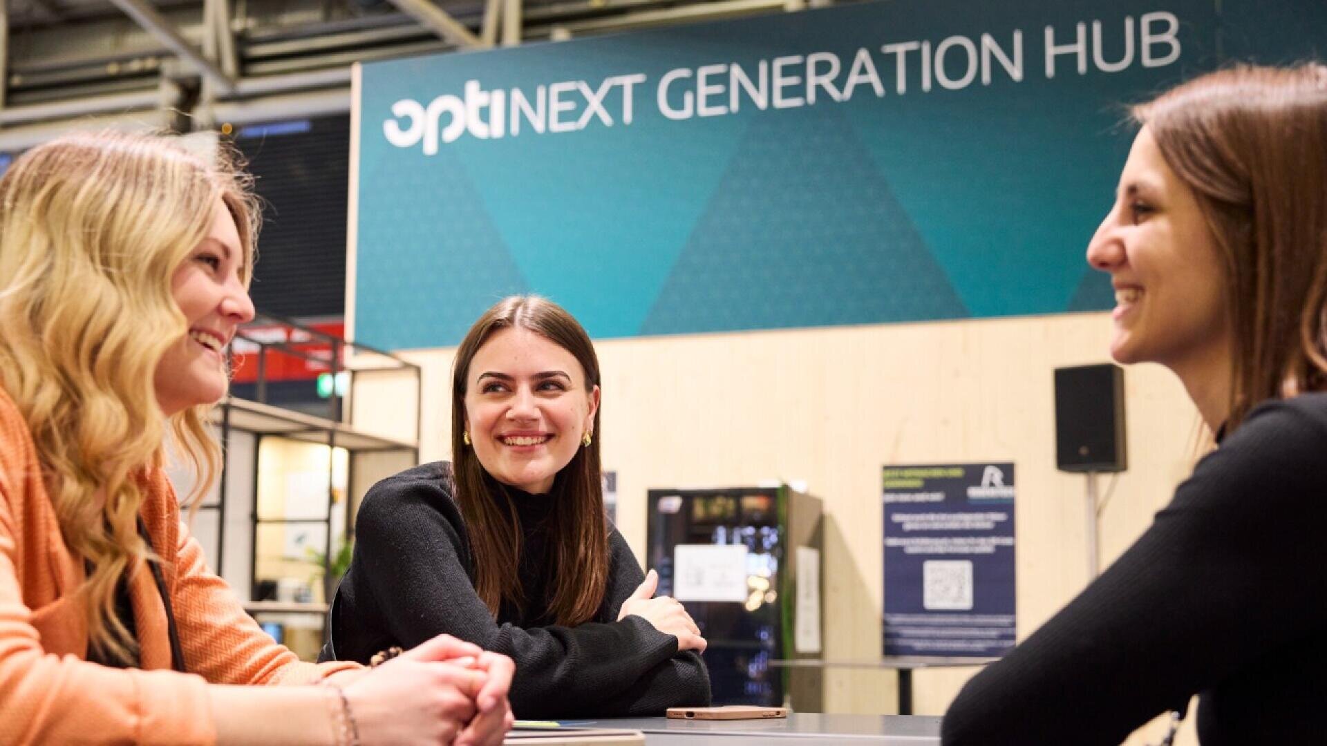 Three women sit at a table, smiling and talking with each other, in front of a sign that reads opti NEXT GENERATION HUB in a modern indoor setting.