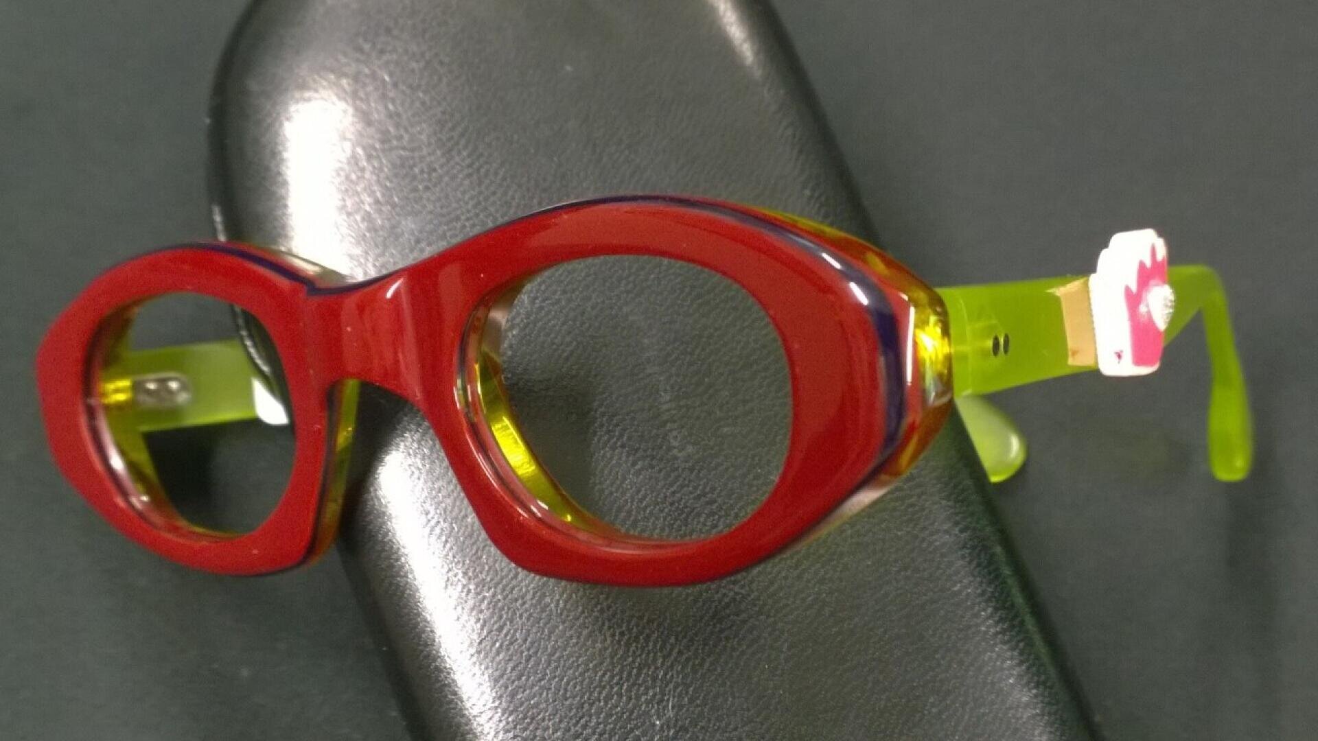A pair of thick, round glasses with red frames and yellow-green arms, resting on a black eyeglass case. A small flower-shaped accessory is attached to one arm.