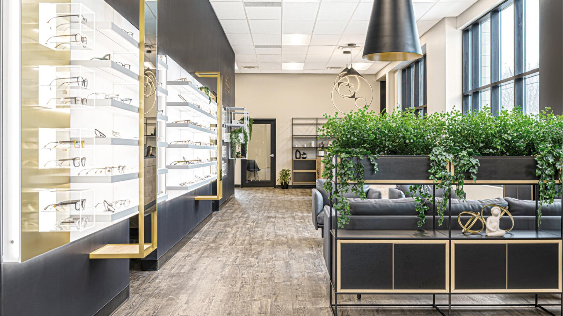 Ein modernes Optikergeschäft mit beleuchteten Wandregalen, großen Fenstern, dekorativen Pflanzen, schwarz-goldenen Akzenten und bequemen Sitzbereichen mit Sofas.