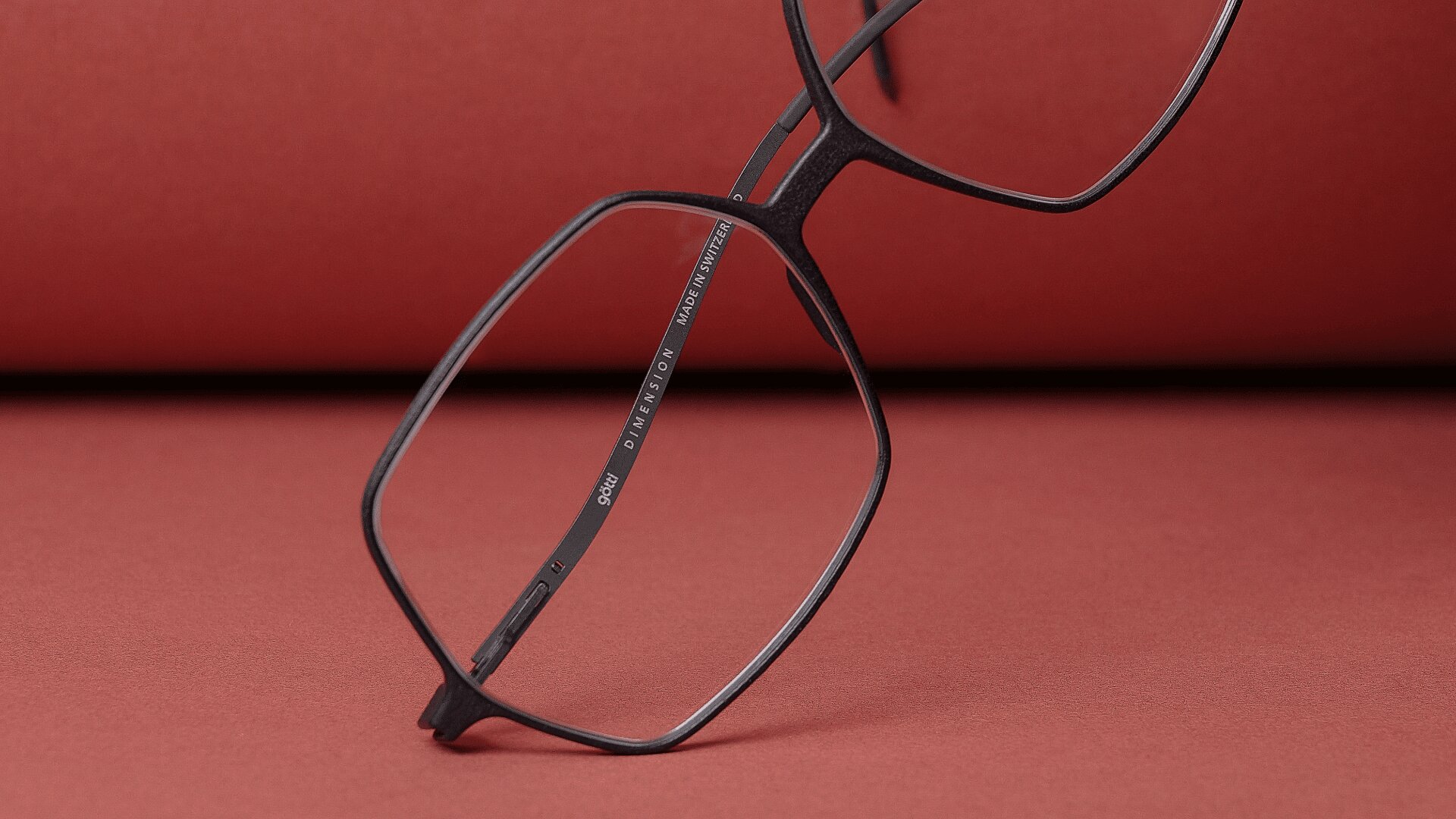 Eine schwarze, quadratisch gerahmte Brille liegt auf einer roten Fläche mit rotem Hintergrund. Die Brille ist hochgeklappt, so dass der dünne Rahmen und eine kleine Inschrift auf der Innenseite eines Bügels zu sehen sind.