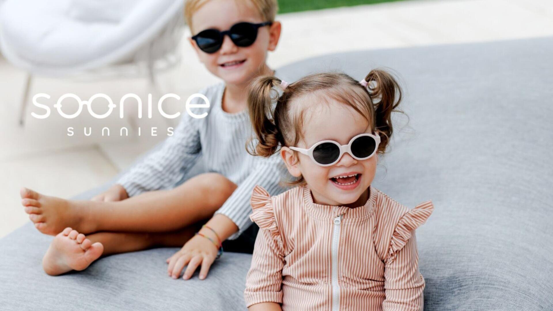 Two young children wearing sunglasses and smiling while sitting outdoors on a light-colored cushion. The child in front has pigtails and wears a striped top, while the child behind wears a light outfit. “SOONICE SUNNIES” text appears on the image.
