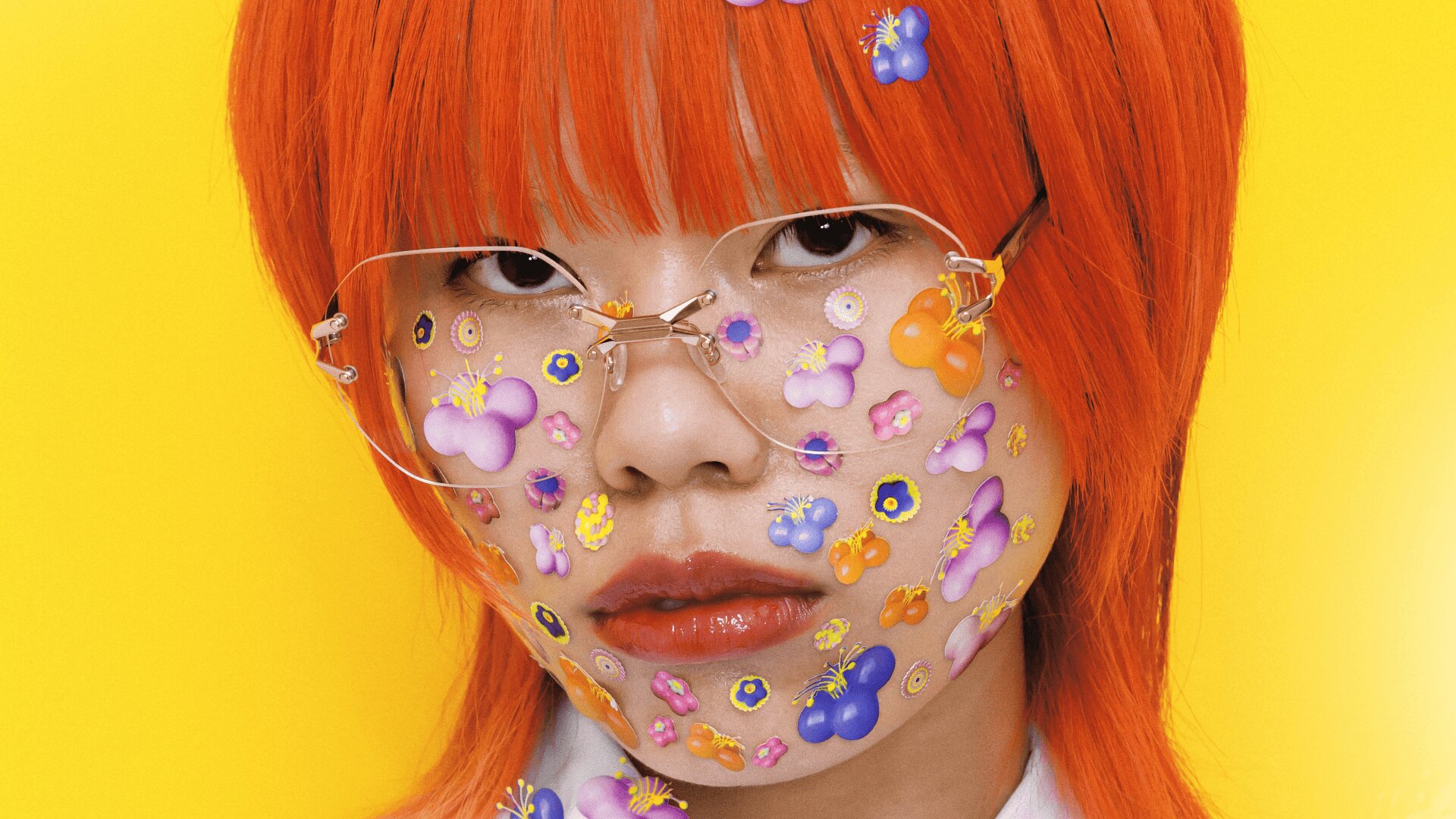 Eine Person mit leuchtend orangefarbenem Haar und einer klaren Brille blickt in die Kamera. Ihr Gesicht ist mit bunten Blumenaufklebern bedeckt. Der Hintergrund ist leuchtend gelb.