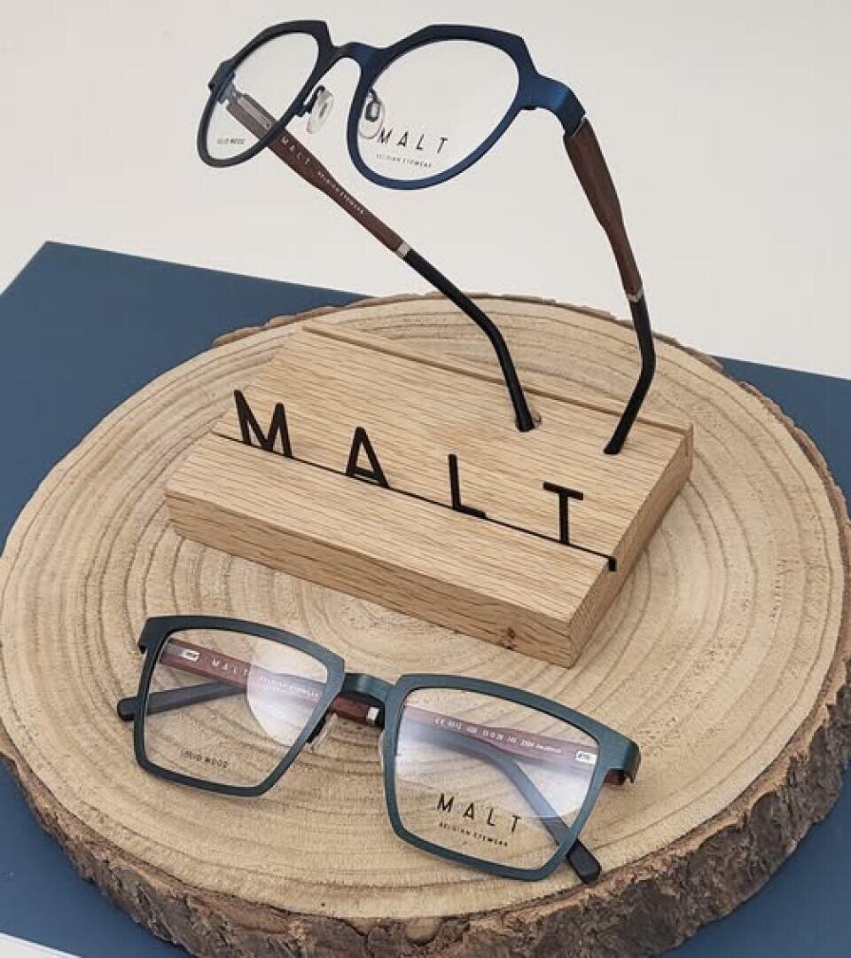 Two pairs of glasses with blue rims are displayed on a round wooden pedestal. One pair of glasses stands upright on a wooden stand with the inscription MALT, while the other pair lies flat in front of it.
