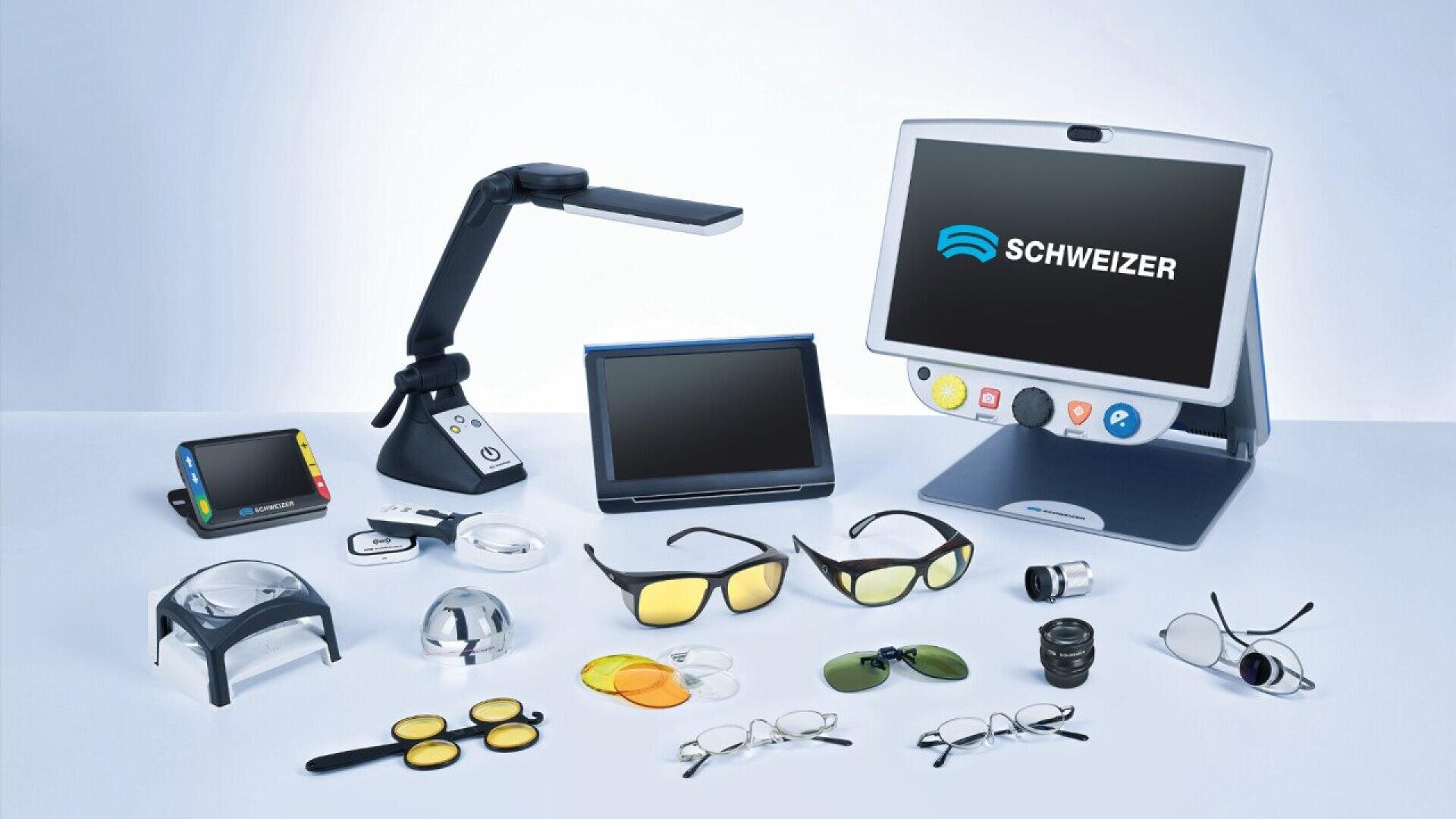 A collection of low vision aids, including electronic magnifiers, screen readers, various magnifying glasses, lenses, and tinted glasses, arranged on a white surface with a SCHWEIZER monitor in the background.