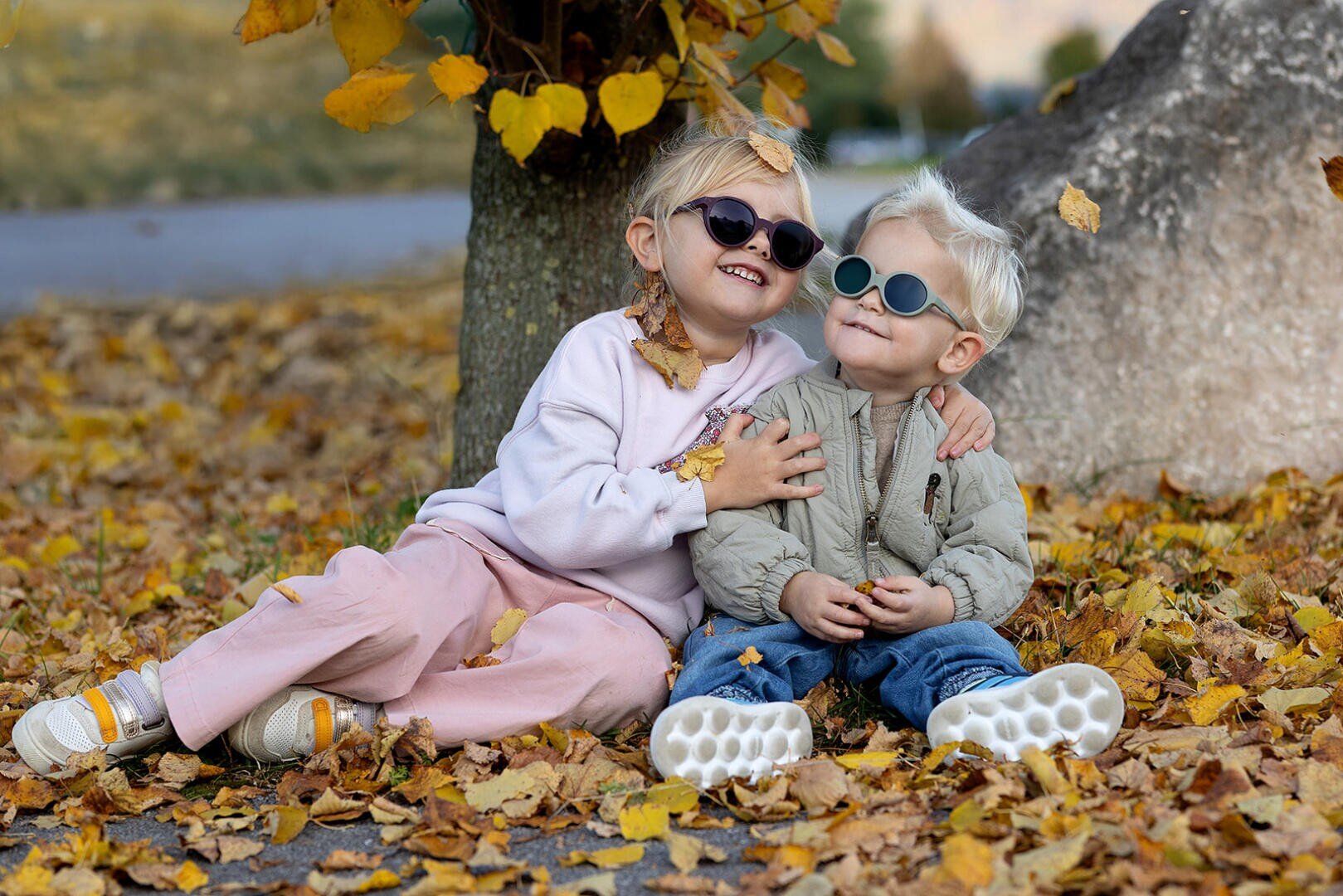 Zwei kleine Kinder mit Sonnenbrillen sitzen auf dem mit Herbstblättern bedeckten Boden, lächeln und umarmen sich in der Nähe eines Baumes. Das eine trägt eine rosa Hose und einen hellen Pullover, das andere eine beige Jacke und eine blaue Hose.