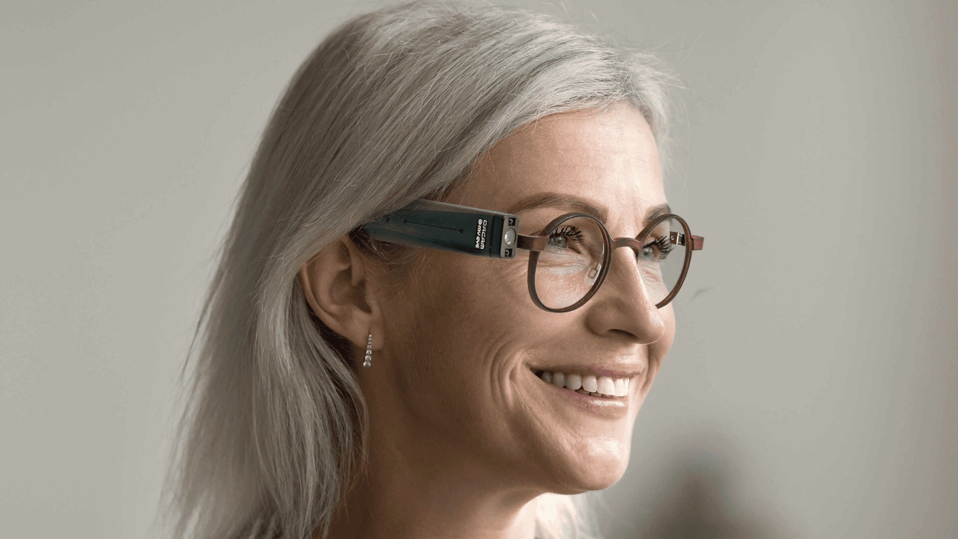 Eine lächelnde ältere Frau mit langen grauen Haaren, einer runden Brille und kleinen Ohrringen ist in Nahaufnahme zu sehen, die vor einem neutralen Hintergrund zur Seite schaut.