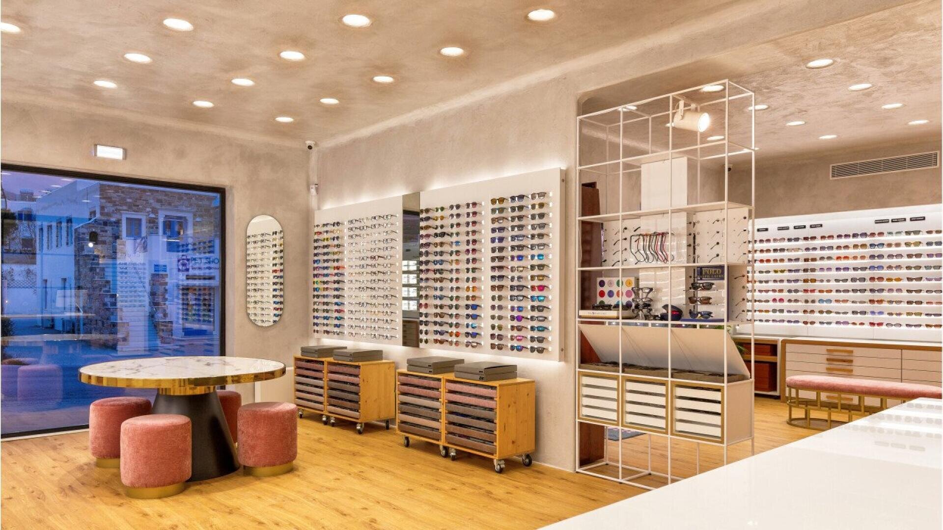 Modern eyewear store with wooden floors, round tables with pink stools, and walls lined with display racks holding many pairs of eyeglasses and sunglasses under bright ceiling lights.