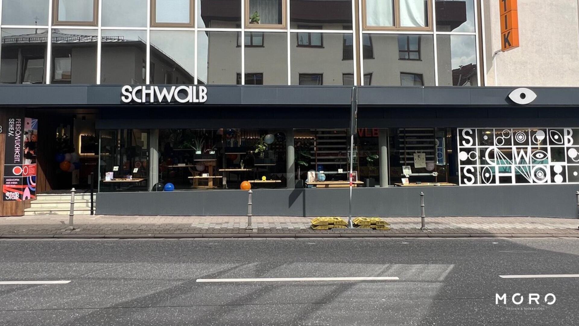 Street view of a modern storefront with large windows displaying interior decor. The sign above the entrance reads SCHWOLB. The pavement and road are visible in the foreground.