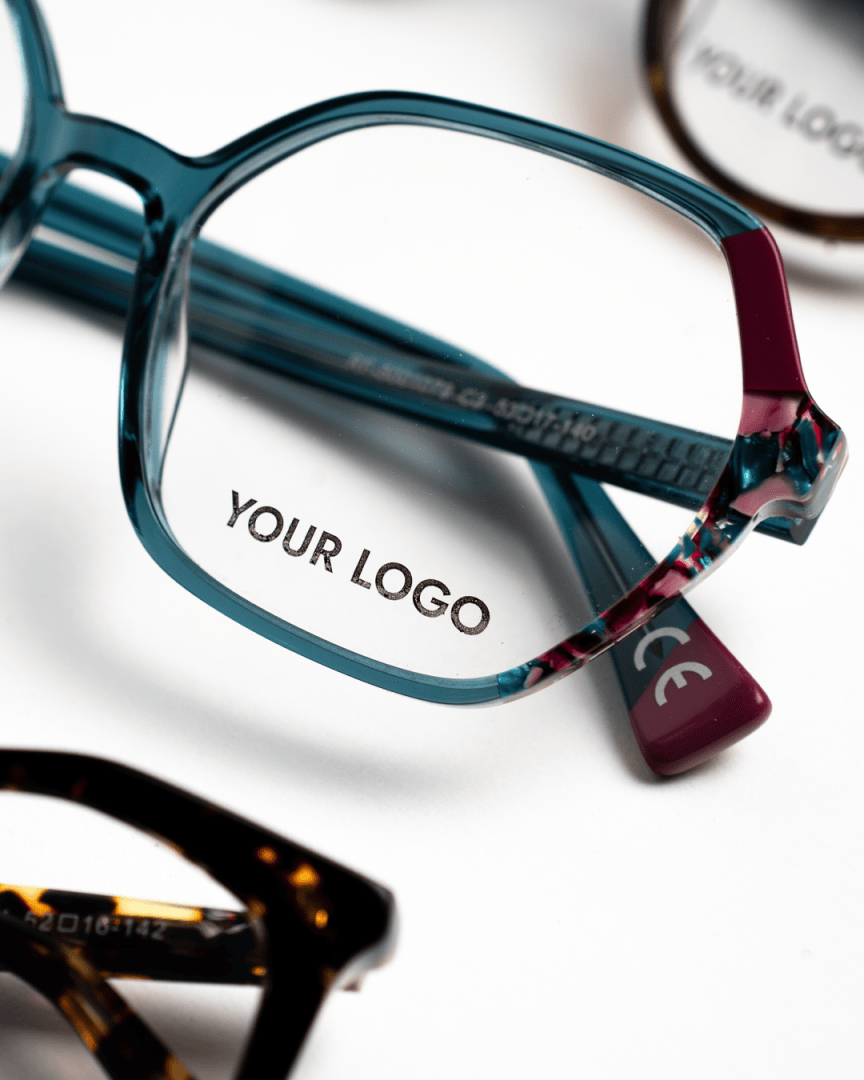 Nahaufnahme eines Brillengestells mit der Aufschrift YOUR LOGO auf dem rechten Glas. Andere Brillen sind teilweise im Hintergrund zu sehen.