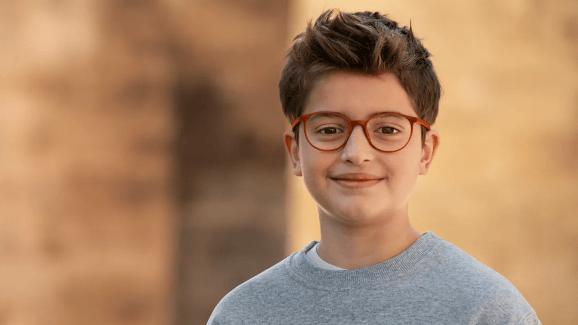 Ein lächelnder Junge mit kurzen braunen Haaren und einer rotgerahmten Brille, der ein graues Sweatshirt trägt, steht im Freien vor einem sanft verschwommenen neutralen Hintergrund.
