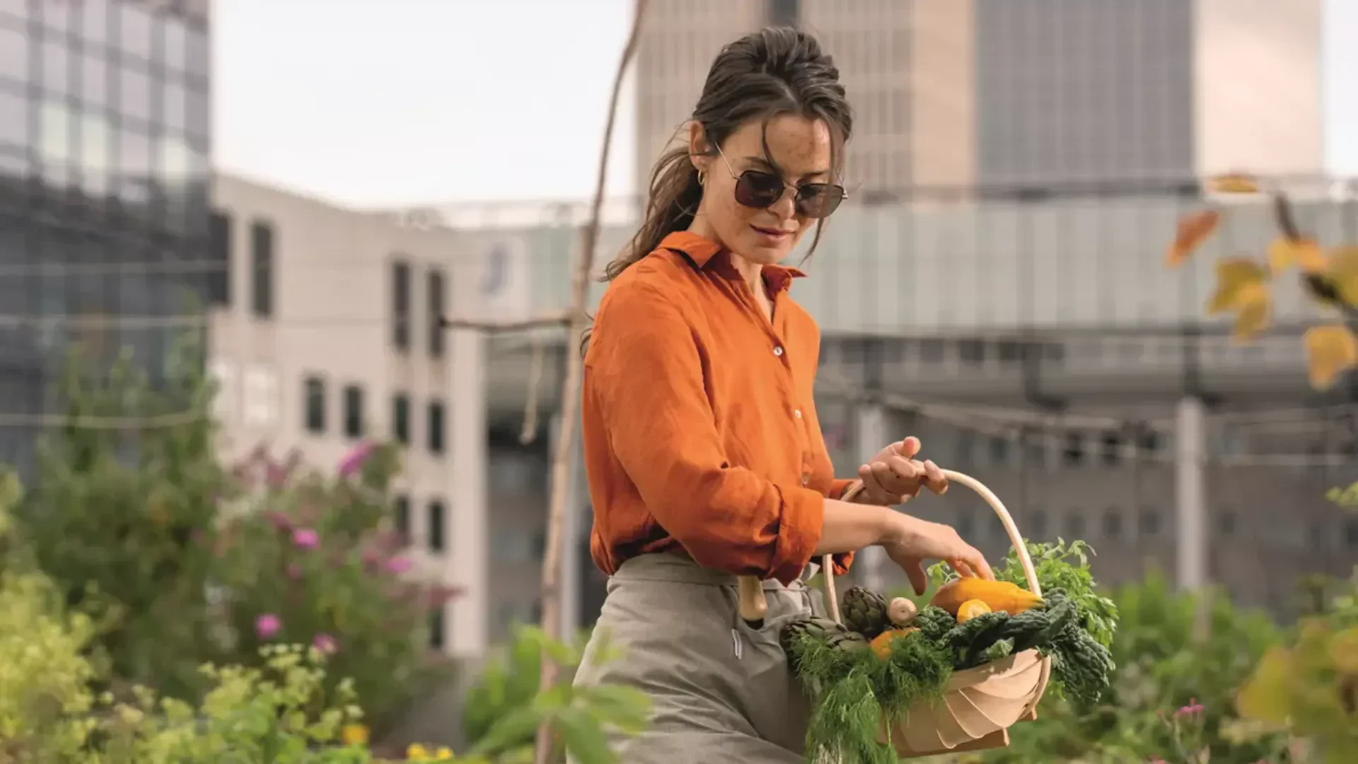 Eine Frau mit Sonnenbrille und orangefarbenem Hemd pflückt Gemüse und Kräuter und legt sie in einen Korb, während sie in einem Dachgarten mit Stadtgebäuden im Hintergrund steht.