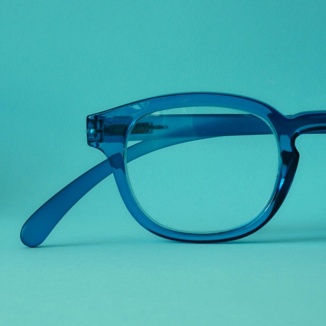 Close-up of the left half of a pair of blue glasses with transparent frames against a matching blue background.