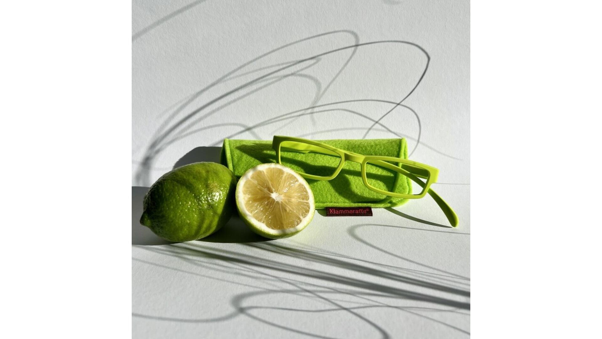 A pair of bright green glasses sits on a green case beside a whole lime and a halved lemon on a white surface. Elegant shadows from the glasses and a plant are cast in the background.