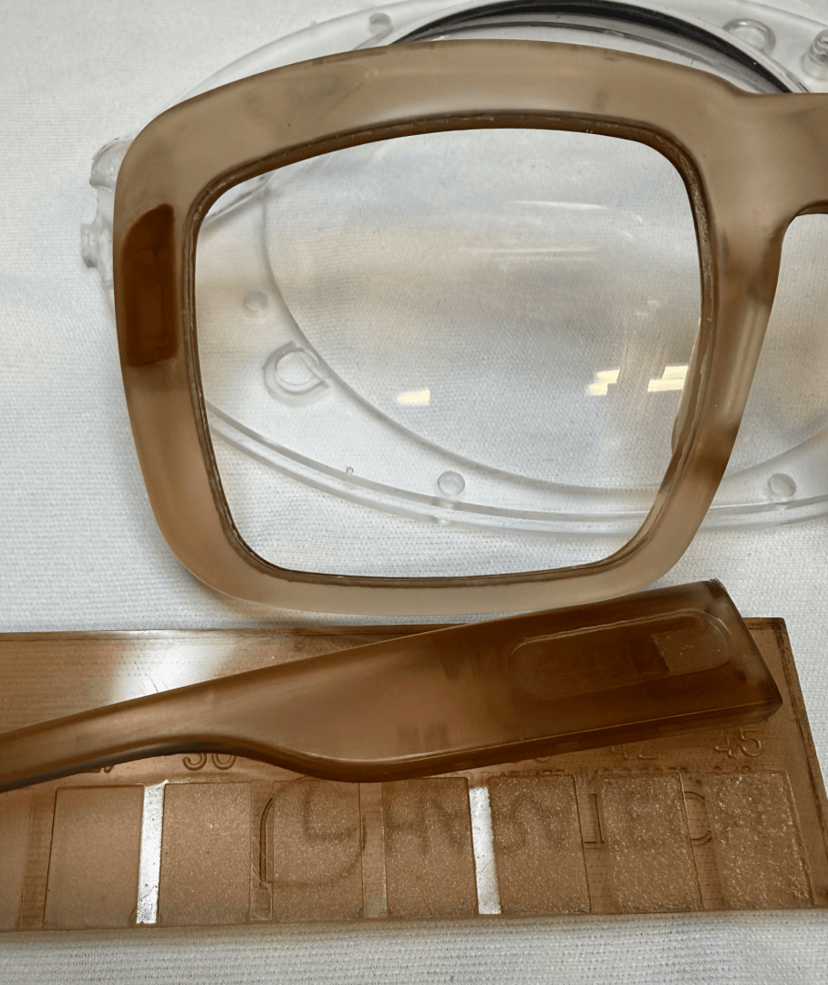 A close-up of a light brown frame with the temple resting on a plastic template or mold showing details of the material and shape.