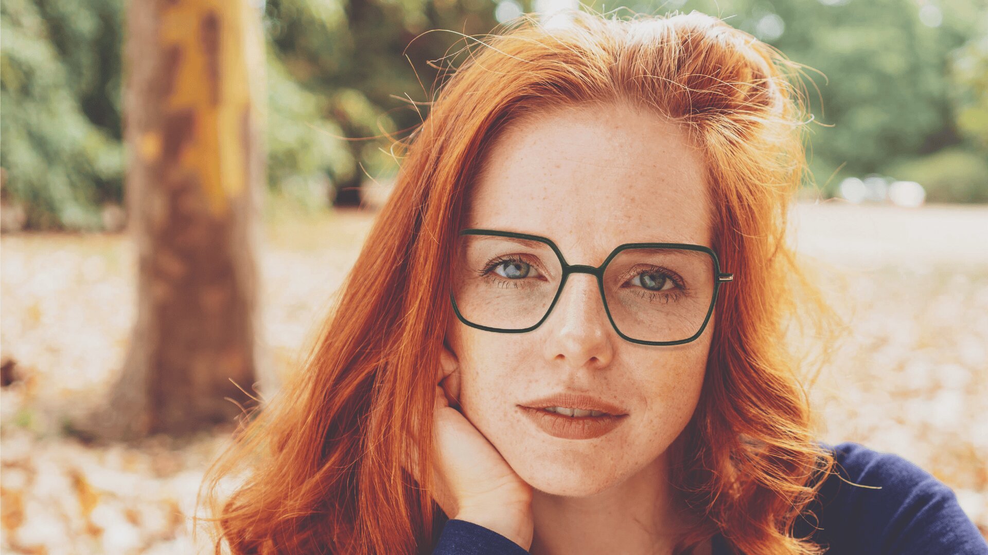 Eine junge Frau mit langen roten Haaren, Sommersprossen und einer großen quadratischen Brille blickt im Freien direkt in die Kamera und stützt ihr Kinn auf ihre Hand. Der Hintergrund ist mit Bäumen und herabgefallenen Blättern unscharf.