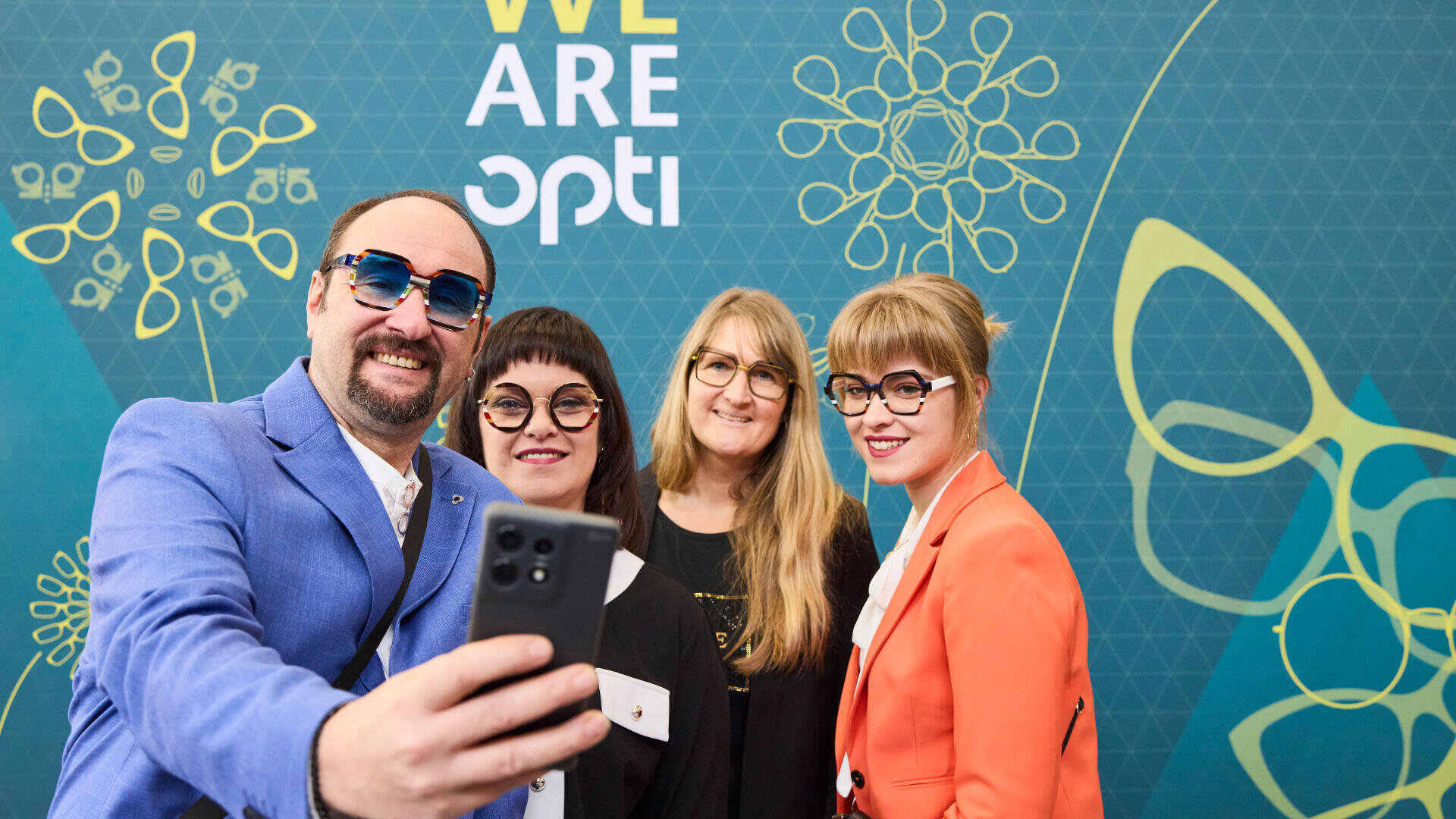 Vier Personen mit Brillen posieren für ein Selfie vor einem blaugrünen Hintergrund mit geometrischen Mustern und dem Schriftzug „WE ARE opti“. Sie lächeln und tragen farbenfrohe, stylische Kleidung.