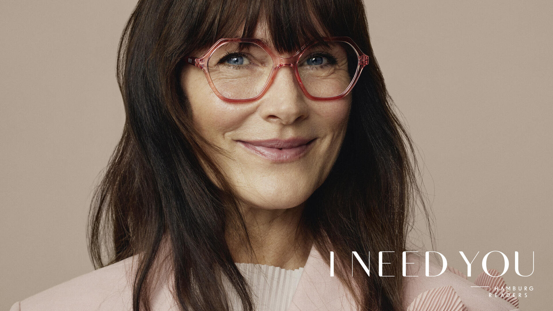 Eine Frau mit langen dunklen Haaren und Pony lächelt sanft. Sie trägt eine rosa Brille mit geometrischem Muster und einen hellrosa Blazer. Der Hintergrund ist beige und in der unteren rechten Ecke steht der Text „I NEED YOU“.