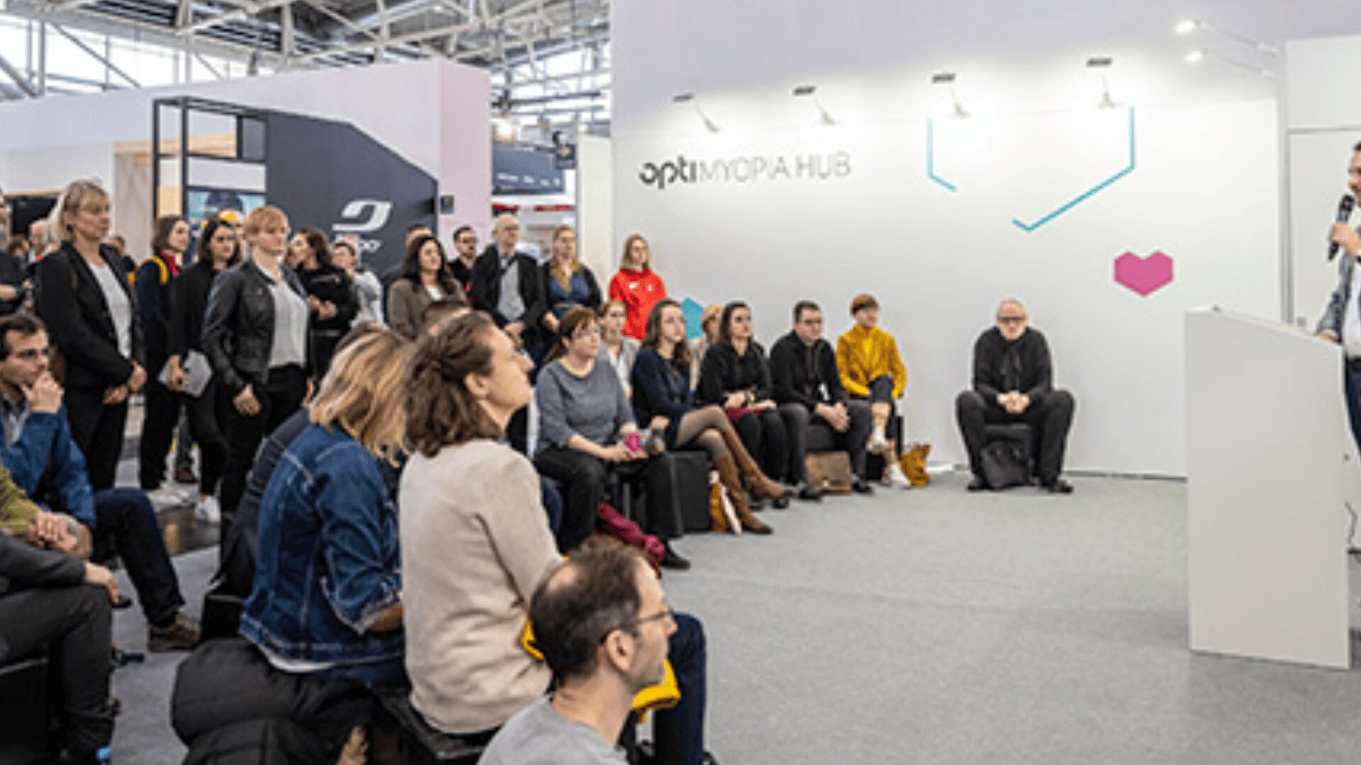 Eine Gruppe von Personen sitzt und steht aufmerksam und hört einem Redner bei einer Veranstaltung oder Konferenz in einem Gebäude zu. Die Umgebung ist modern eingerichtet, mit einer weißen Wand mit der Aufschrift OPTIK und FORA HUB im Hintergrund.