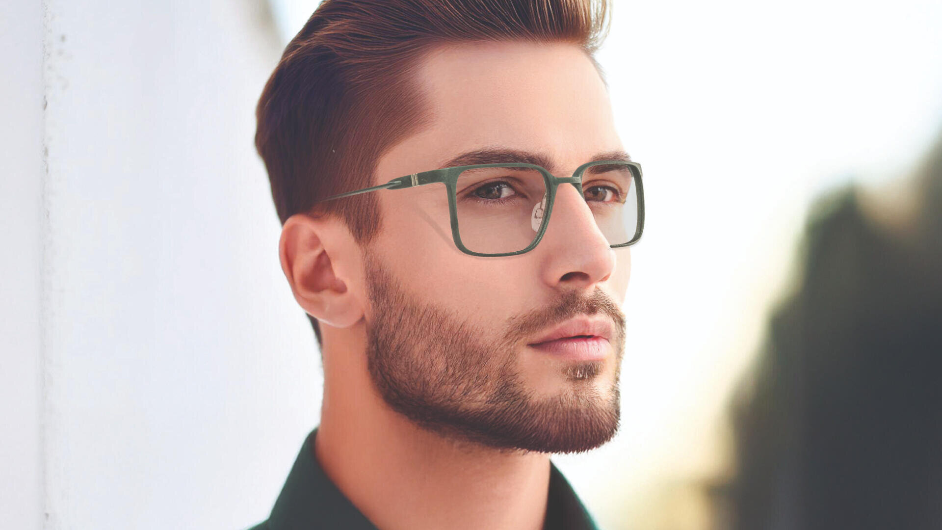 Ein Mann mit kurzen braunen Haaren, gestutztem Bart und Brille blickt nachdenklich in die Ferne. Er trägt ein dunkelgrünes Hemd mit Kragen und steht neben einer weißen Wand mit unscharfem Außenhintergrund.