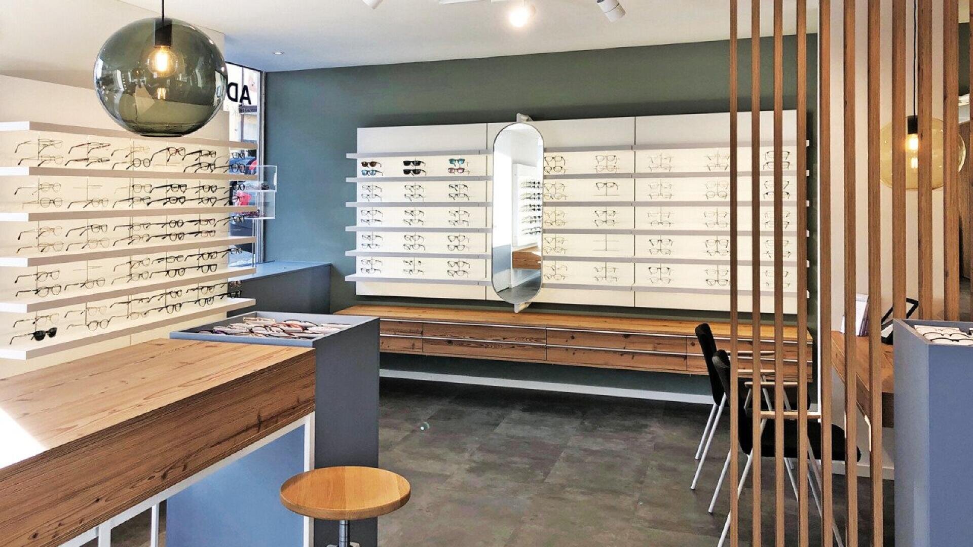 A modern eyewear store interior with rows of eyeglasses displayed on white shelves, a central wooden table, chairs, a hanging glass light, and a tall mirror on the back wall.