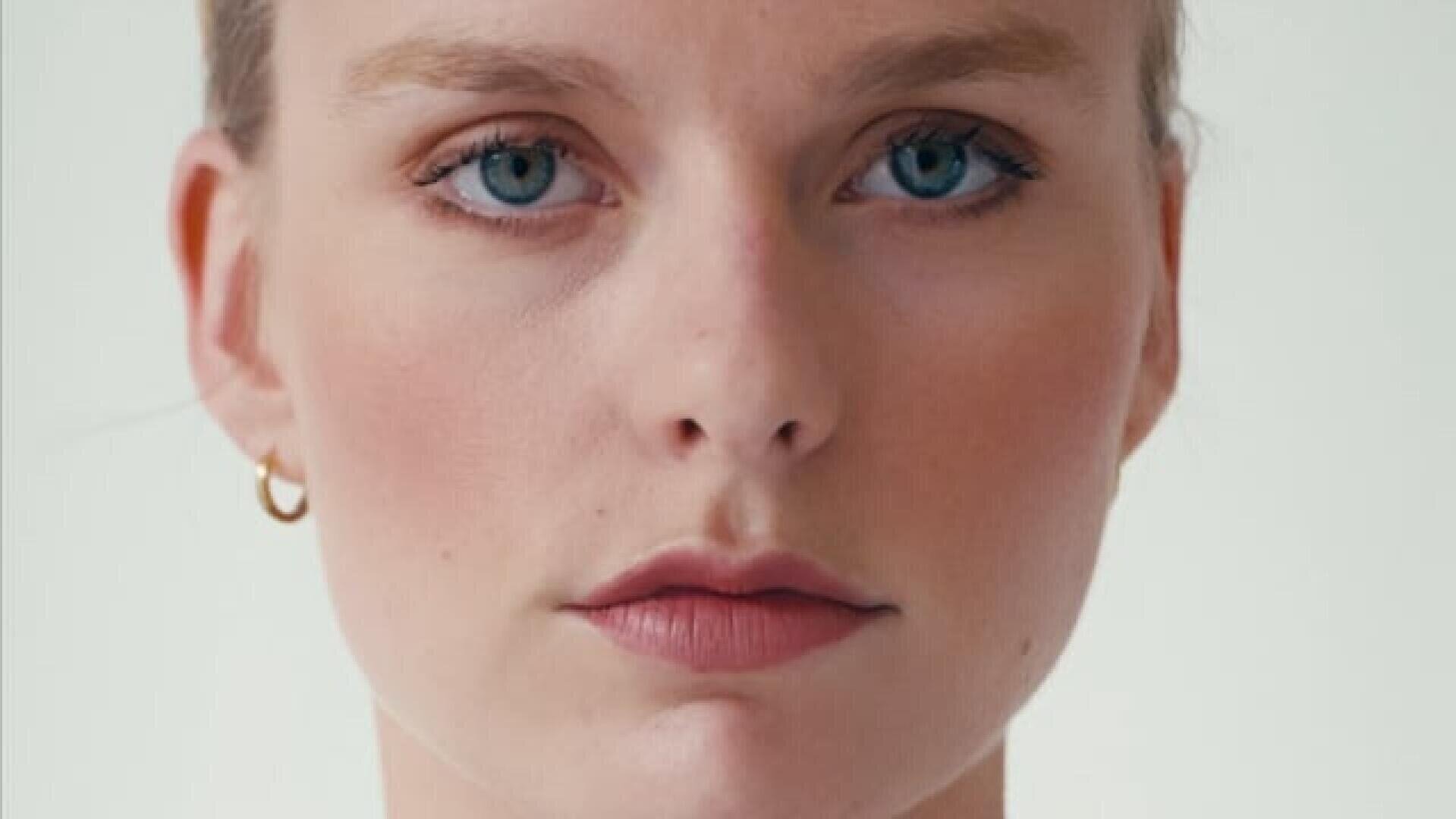 Nahaufnahme eines Porträts einer Person mit heller Haut, blauen Augen und hellrosa Lippen, die einen kleinen goldenen Ohrring trägt und mit neutralem Gesichtsausdruck direkt in die Kamera schaut, vor einem schlichten Hintergrund.
