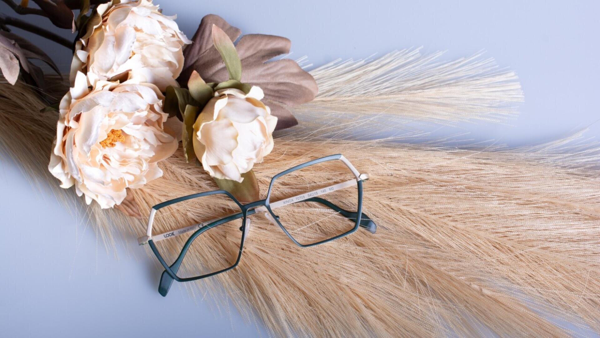 Eine Brille mit quadratischem Rahmen liegt auf beigem, getrocknetem Pampasgras neben hellcremefarbenen Kunstblumen vor einem sanften blauen Hintergrund.