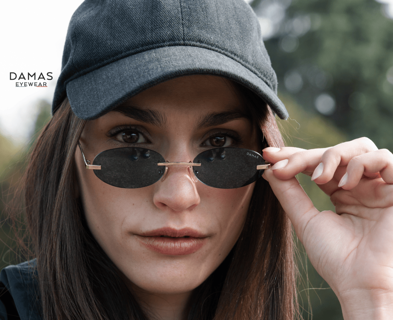 A woman wearing a black cap and oval sunglasses looks directly at the camera, holding her sunglasses with one hand. The DAMAS EYEWEAR logo appears in the top left corner. Trees are blurred in the background.