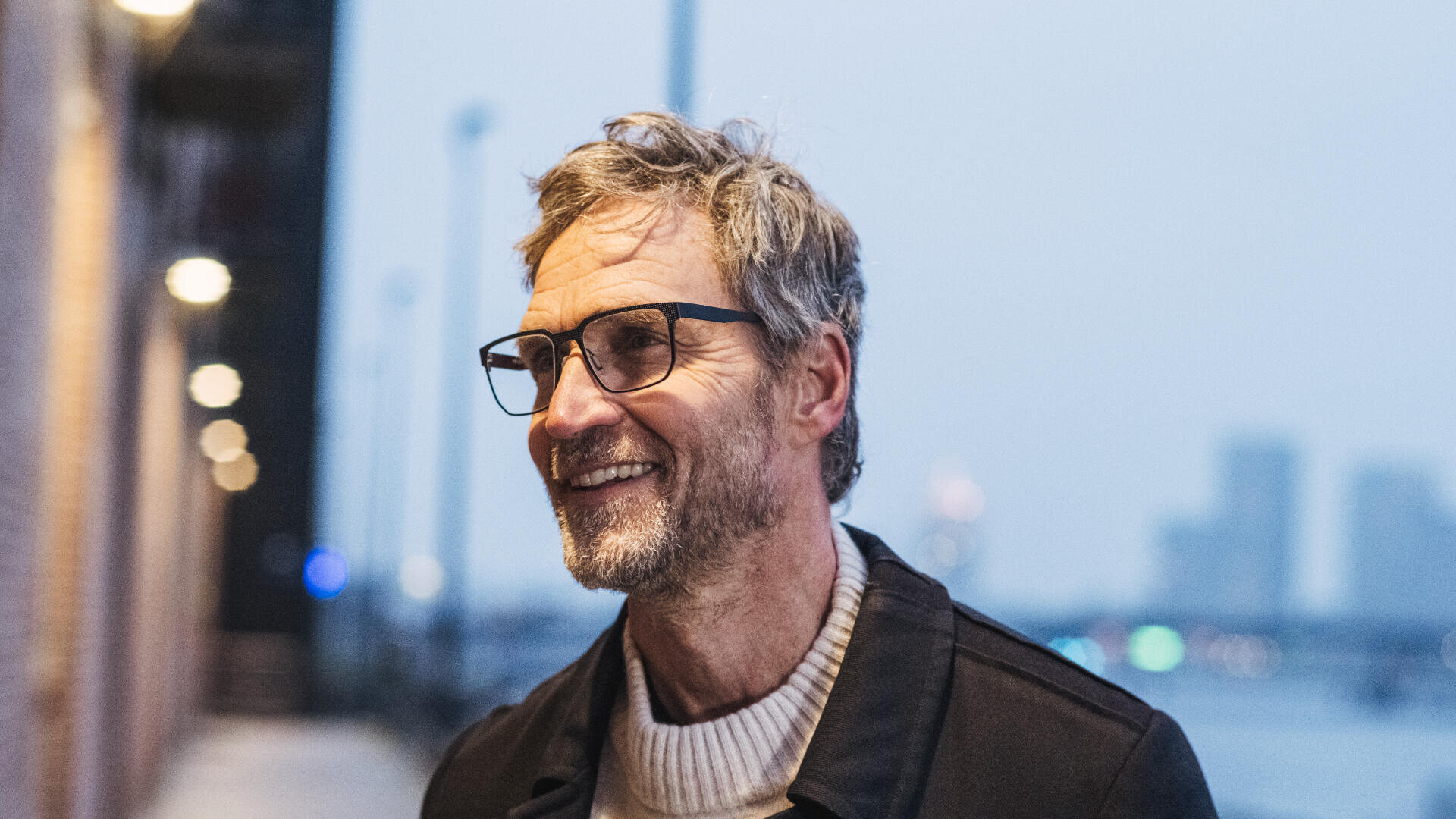 Ein Mann mittleren Alters mit grauem Haar, Brille und Bart steht an einem bewölkten Tag lächelnd im Freien in der Nähe einer Uferpromenade und trägt einen hellen Pullover und eine dunkle Jacke.