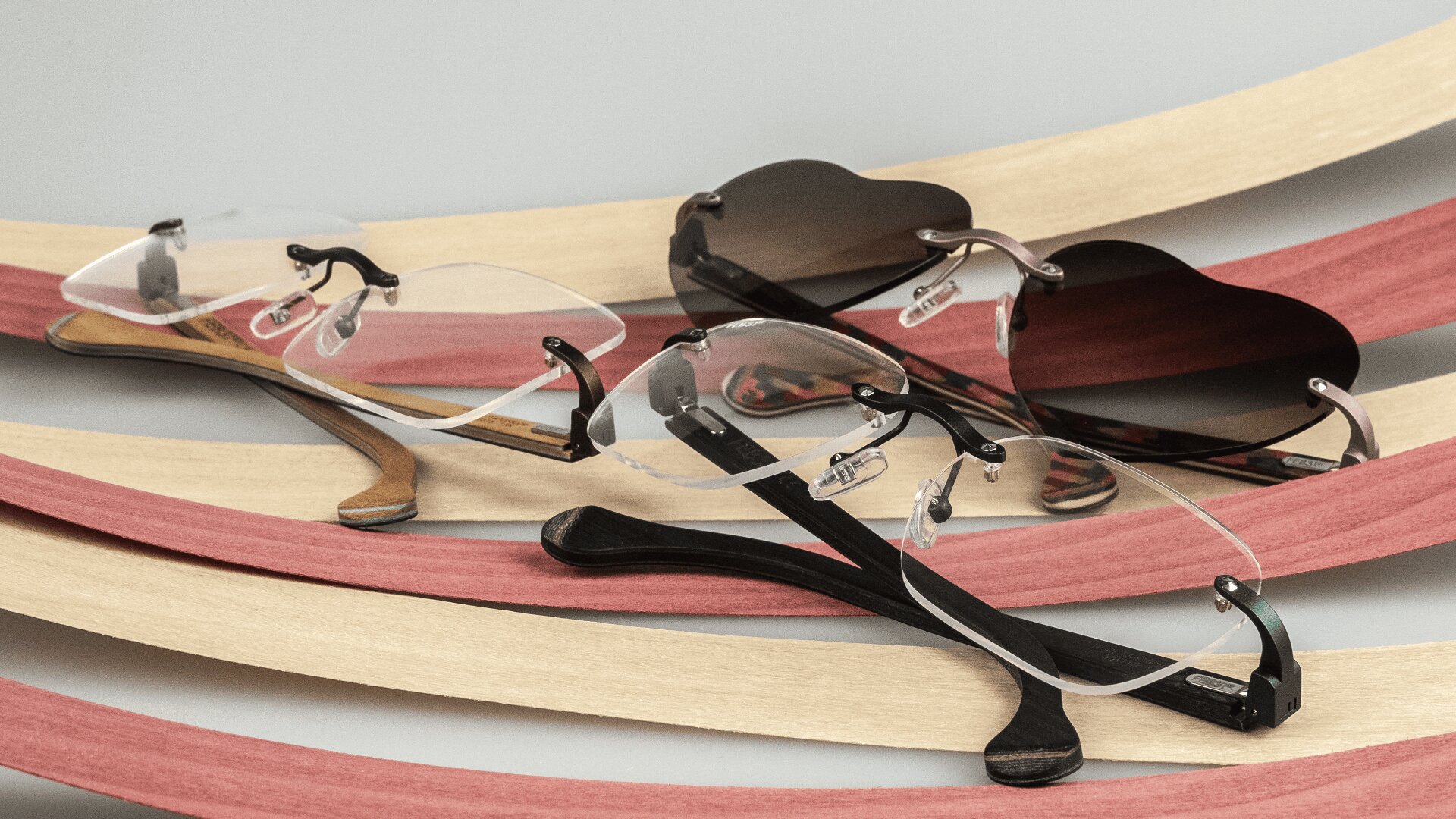 Zwei randlose Brillen, eine mit klaren und eine mit dunkel getönten Gläsern, ruhen auf geschwungenen Holzlatten in Beige- und Rottönen. Die Brillen haben minimalistische Details aus Metall und Holz.