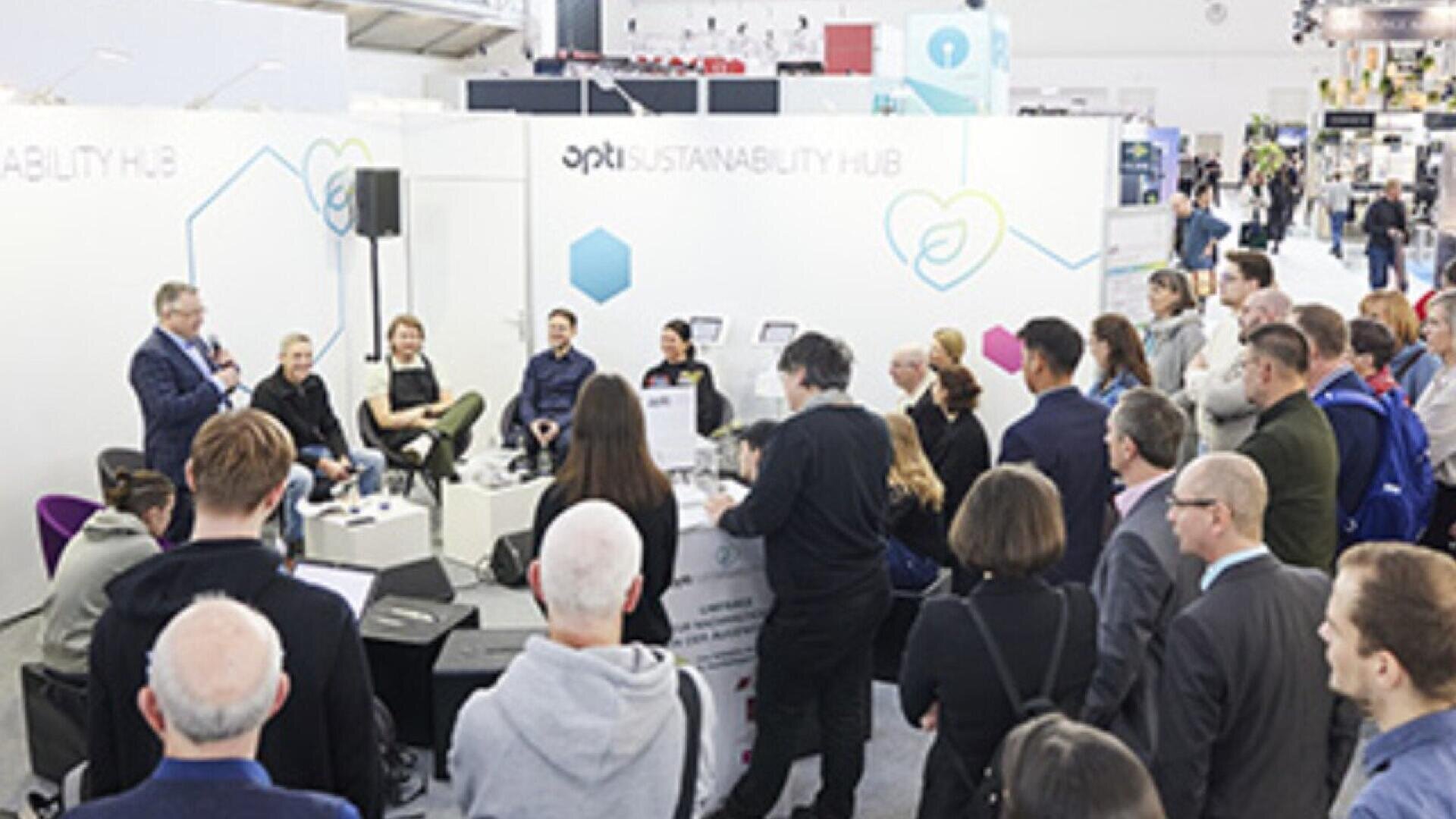 A group of people attend a panel discussion at a trade show or conference, seated and standing around speakers in front of a white booth labeled Sustainability Hub.