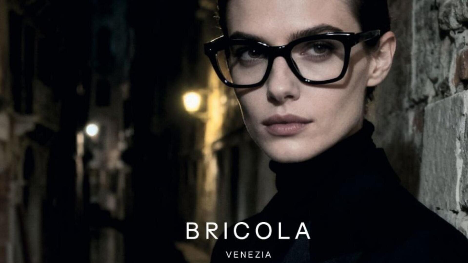 Eine Person mit großer schwarzer Brille und schwarzem Outfit steht in einer nächtlichen, schwach beleuchteten Gasse. Am unteren Rand des Bildes erscheint der Text BRICOLA VENEZIA.