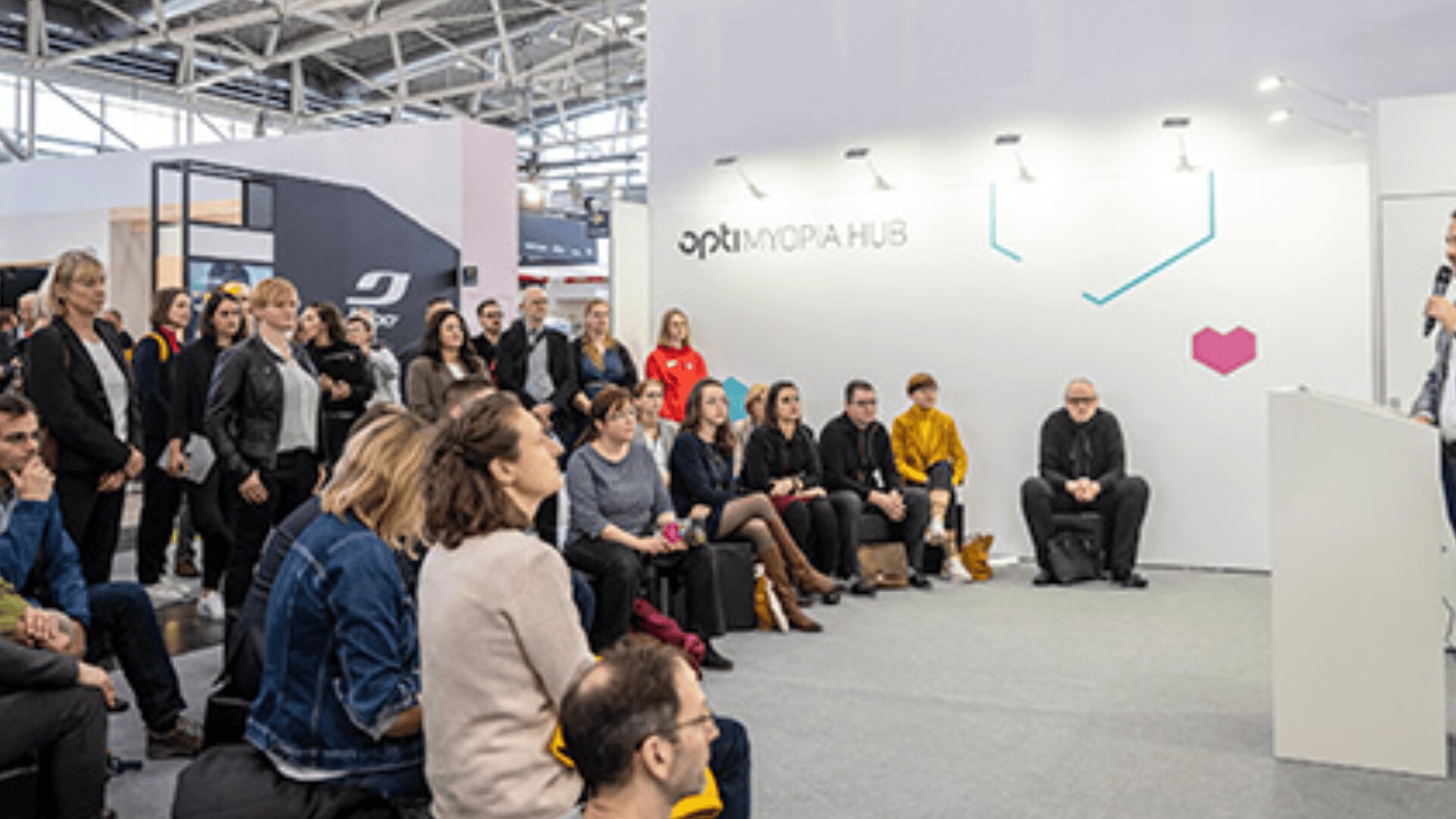 Eine Gruppe von Menschen sitzt und steht aufmerksam da und hört einem Redner bei einer Veranstaltung oder Konferenz in einer Halle zu. Im Hintergrund ist eine weiße Wand mit der Aufschrift Opti MORPH HUB zu sehen.