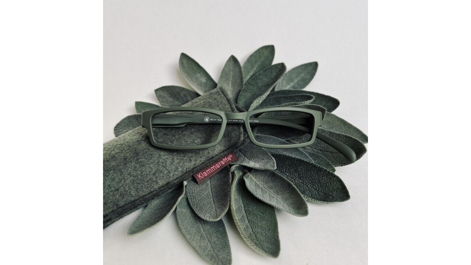 Rectangular green eyeglasses rest on sage leaves and a matching green felt case, with a maroon tag labeled Mamamatie visible on the case.